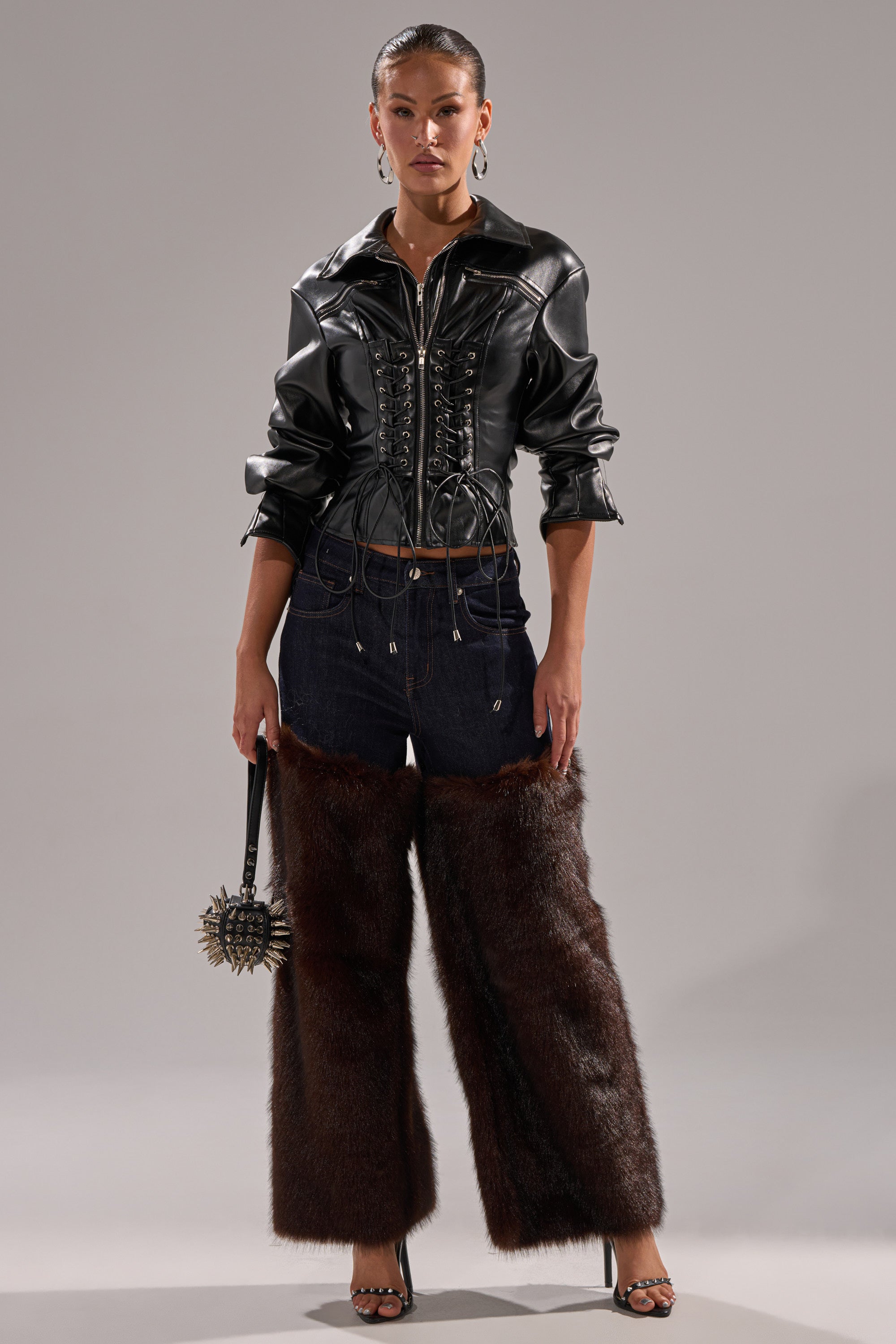 A woman faces forward in the AUDREY FAUX LEATHER BLOUSE, styled like a black lace-up moto jacket, paired with dark denim pants featuring brown faux fur legs, high heels, and a spiked handbag against a plain gray background.