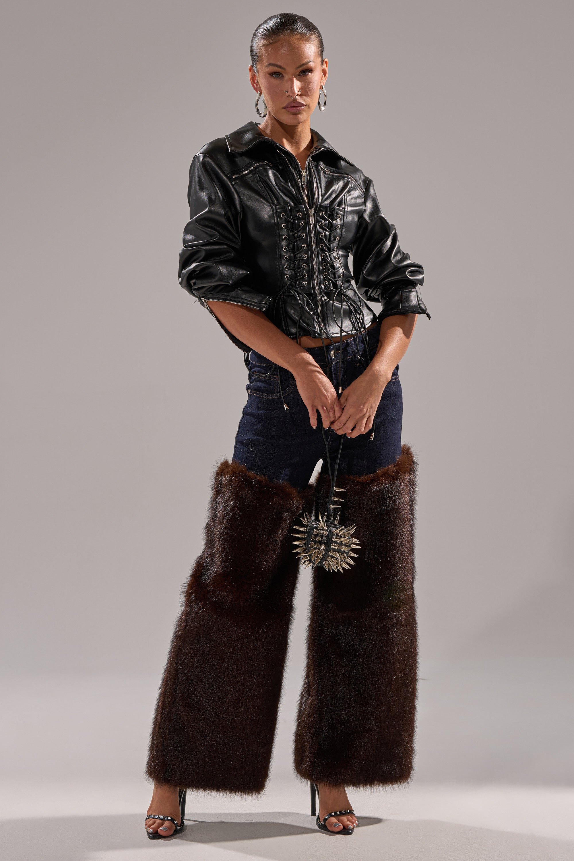 A woman stands confidently in the AUDREY FAUX LEATHER BLOUSE, dark pants, and oversized brown furry chaps over high heels, holding a sun-shaped clutch, with slicked-back hair and large hoop earrings.
