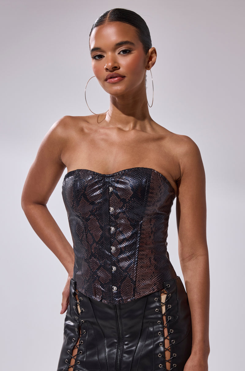 A woman with slicked-back hair and large hoop earrings wears the TIFFANY SNAKE CORSET, a strapless faux snakeskin leather top with metal buttons, paired with a black leather skirt featuring lace-up details. She poses confidently against a plain background. 0