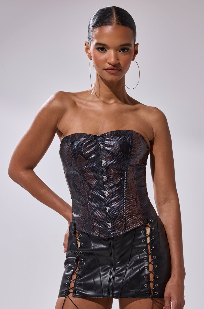 A woman with slicked-back hair confidently models the TIFFANY SNAKE CORSET—a strapless faux snakeskin leather top—paired with a black leather lace-up mini skirt, posing against a plain light background. 1