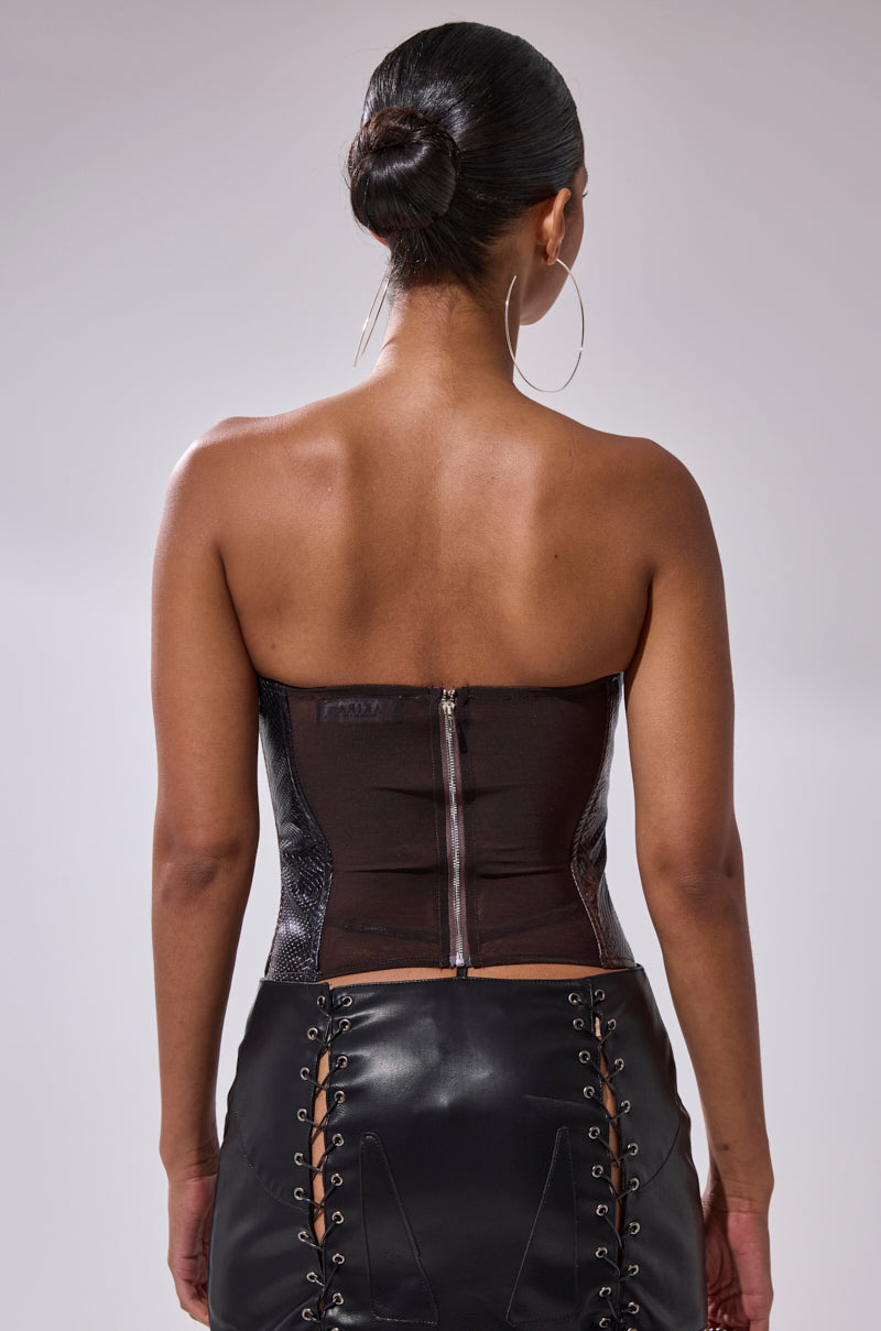 A woman with her hair in a bun, wearing large hoop earrings and the TIFFANY SNAKE CORSET—a strapless faux snakeskin leather top with a back zipper—and a black lace-up faux leather skirt, is shown from behind. 3