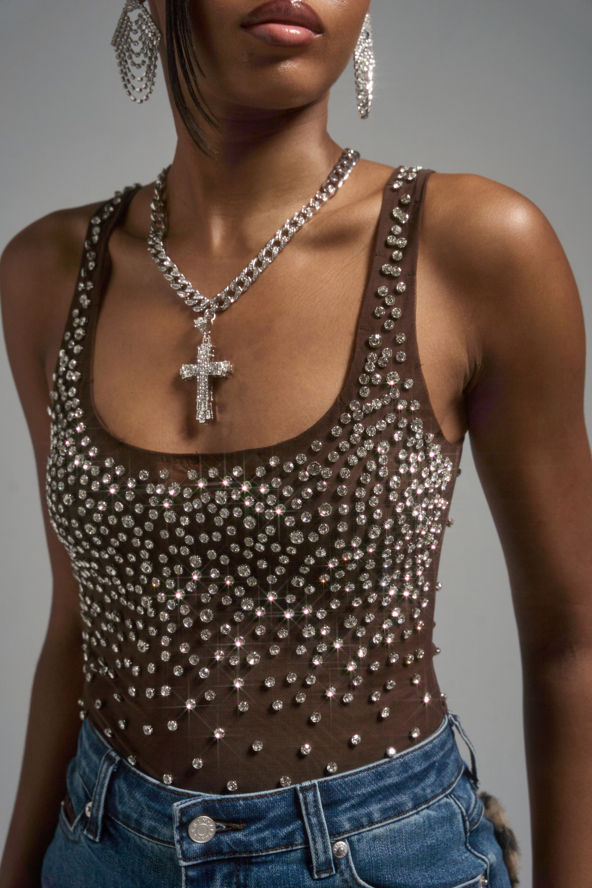 A woman wears the POP IT OFF QUEEN RHINESTONE BODYSUIT from AKIRA Label with blue jeans, large silver earrings, and a chunky chain cross necklace. The photo is cropped at her shoulders and waist.
