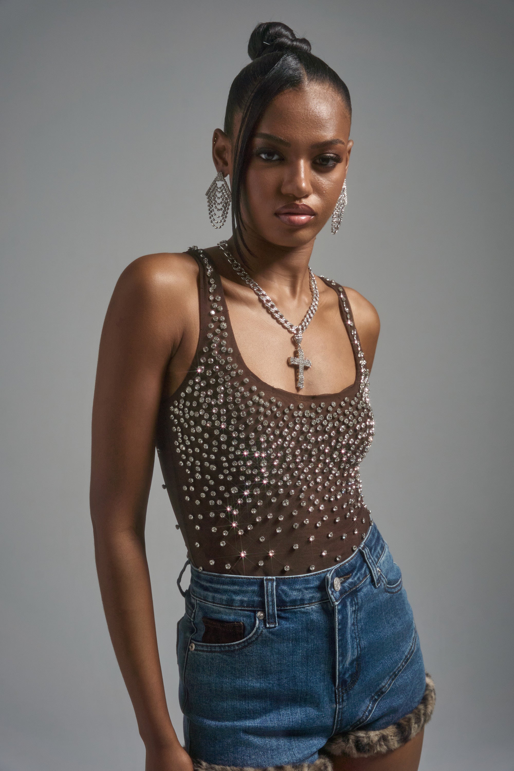 A woman wearing the POP IT OFF QUEEN RHINESTONE BODYSUIT, denim shorts with fur trim, and silver jewelry stands against a plain gray background, looking confidently at the camera.