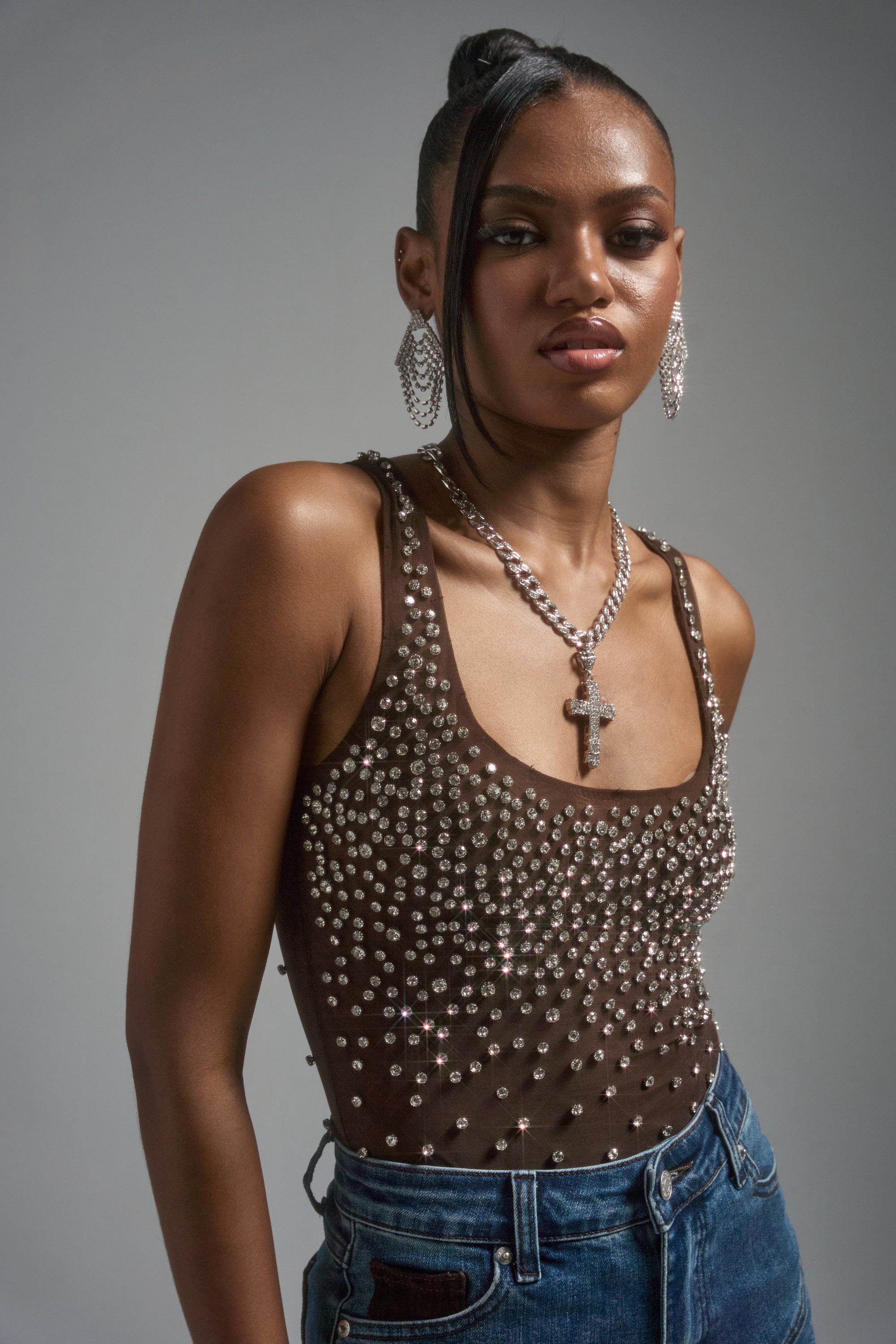 A woman models the POP IT OFF QUEEN RHINESTONE BODYSUIT with blue jeans, accessorized with dangling earrings and a cross necklace. Her hair is in a sleek updo with a side part.