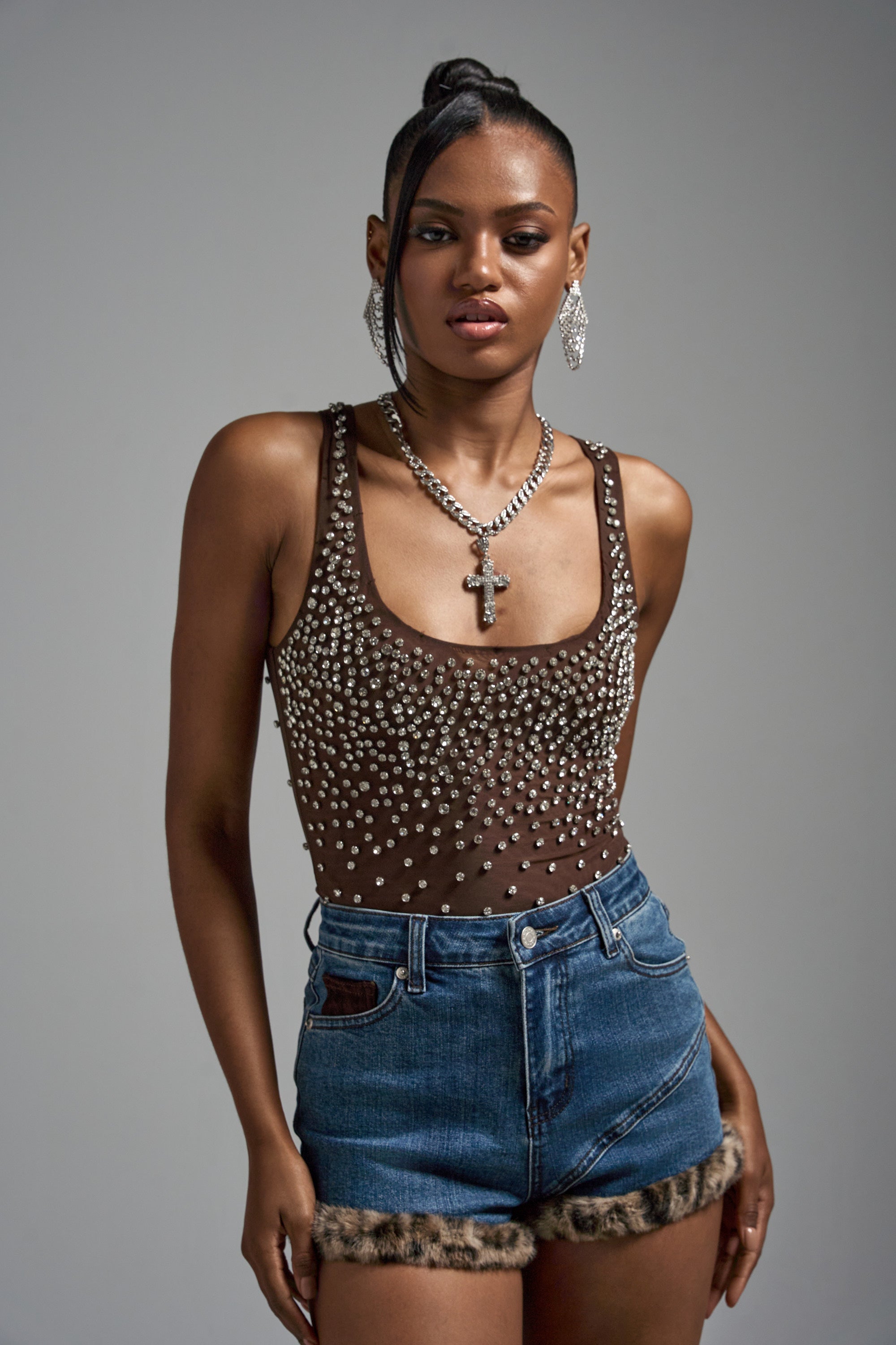 A woman poses confidently against a plain background wearing the POP IT OFF QUEEN RHINESTONE BODYSUIT, denim shorts with faux fur trim, a cross necklace, large earrings, and her hair styled up.