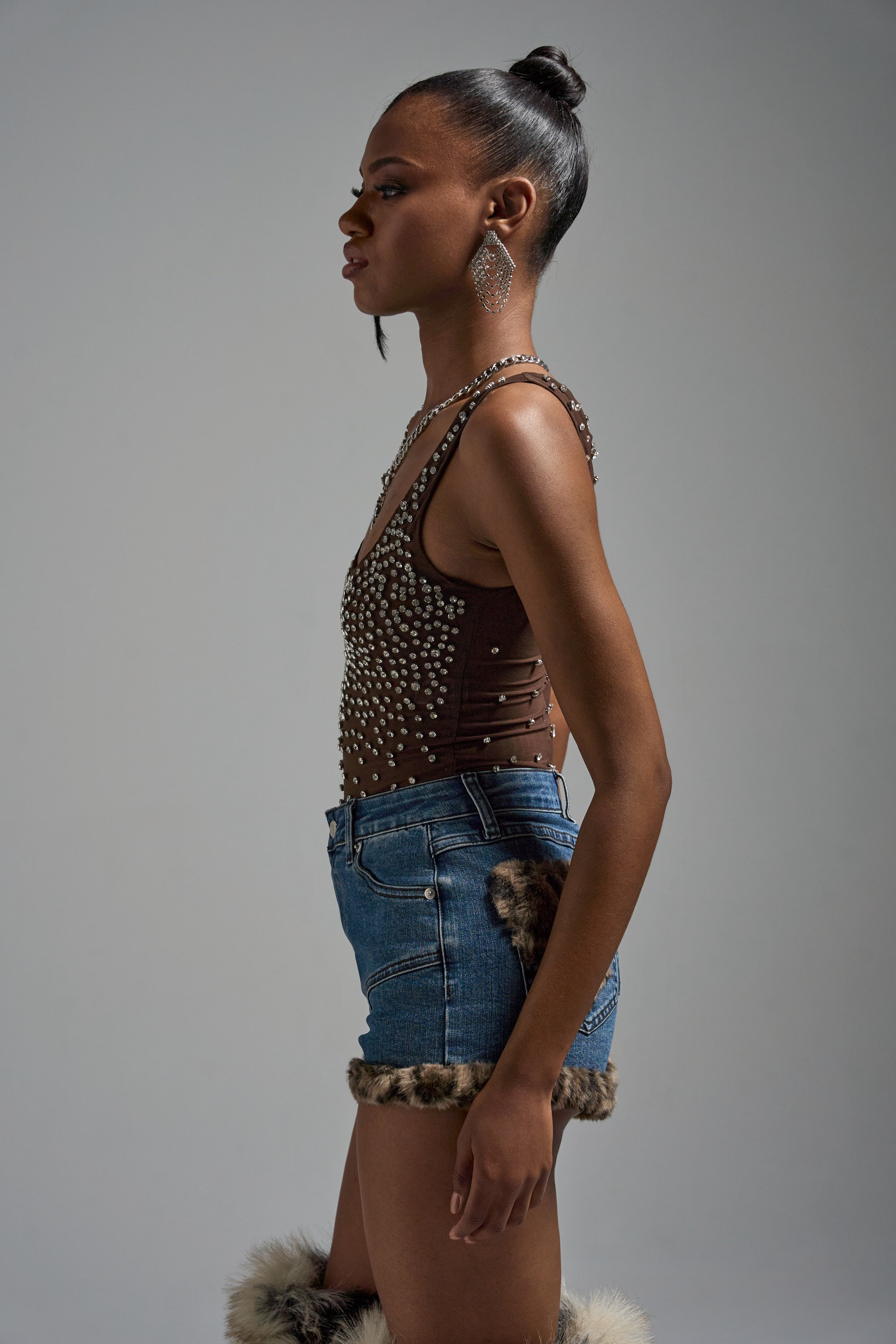 A woman stands in profile against a neutral background, wearing the POP IT OFF QUEEN RHINESTONE BODYSUIT, denim shorts with faux fur trim, large dangling earrings, and her hair styled in a high bun.