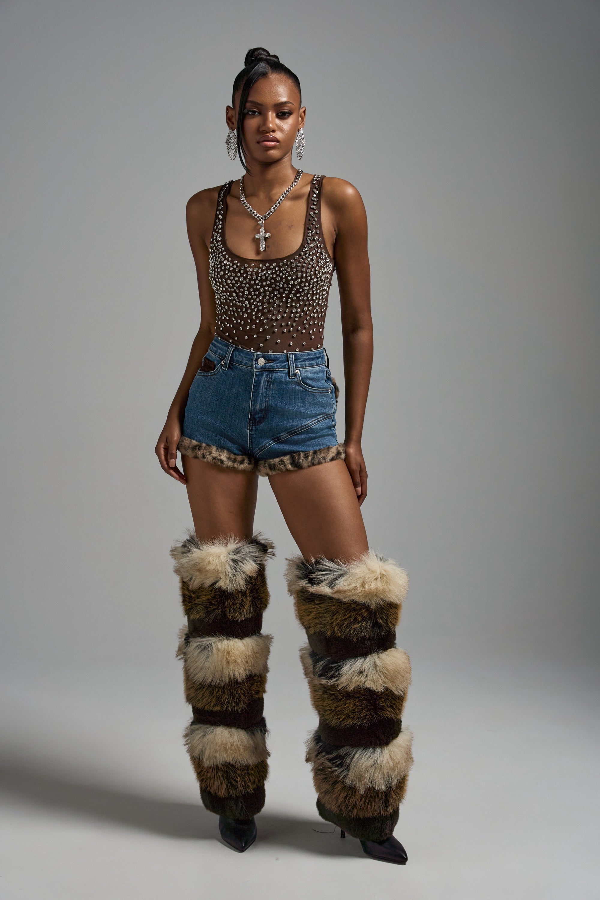A woman poses against a plain background wearing the POP IT OFF QUEEN RHINESTONE BODYSUIT, denim shorts with fur trim, tall furry brown and beige leg warmers, hoop earrings, layered necklaces, and black heels.