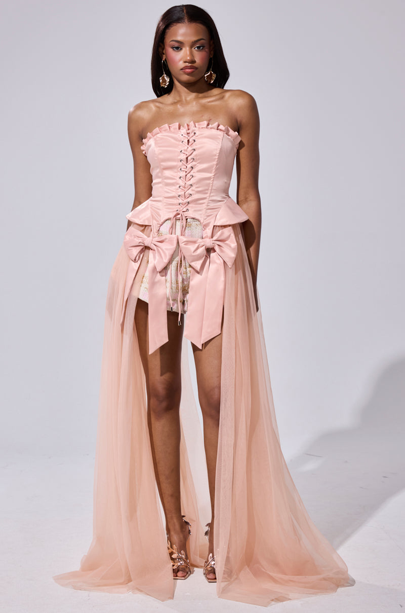A woman wears the HIGH TEA TULLE TRIM CORSET, a strapless blush pink dress featuring a satin corset bodice, large waist bows, and a sheer floor-length tulle overlay atop short underlayers. She stands facing forward on a plain background.