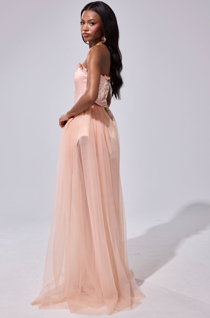 A woman stands in profile, wearing the HIGH TEA TULLE TRIM CORSET—a peach tulle gown with a fitted corset bodice and flowing skirt. Her long dark hair frames her confident pose against a light background.