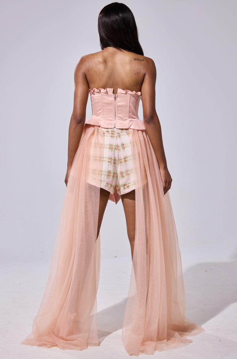 A woman faces away in the HIGH TEA TULLE TRIM CORSET—a strapless peach satin top with zipper and sheer tulle train—paired with high-waisted white and gold patterned shorts, set against a plain light backdrop.