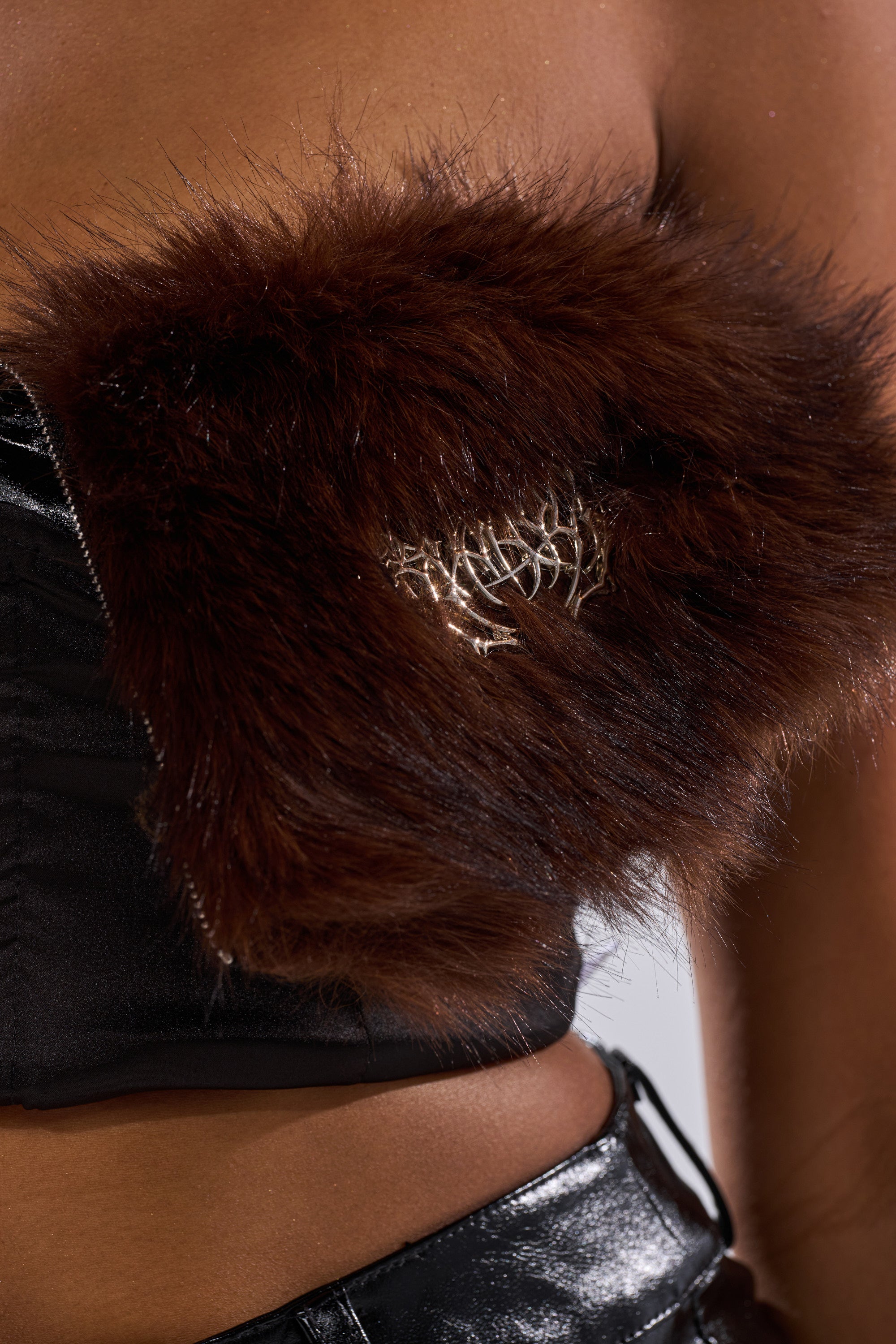 A close-up of a person in a black shiny outfit styled with the ALL THE BIG ONES CORSET WITH FUR, featuring a metallic decorative piece on the chest and visible skin above the waistband.