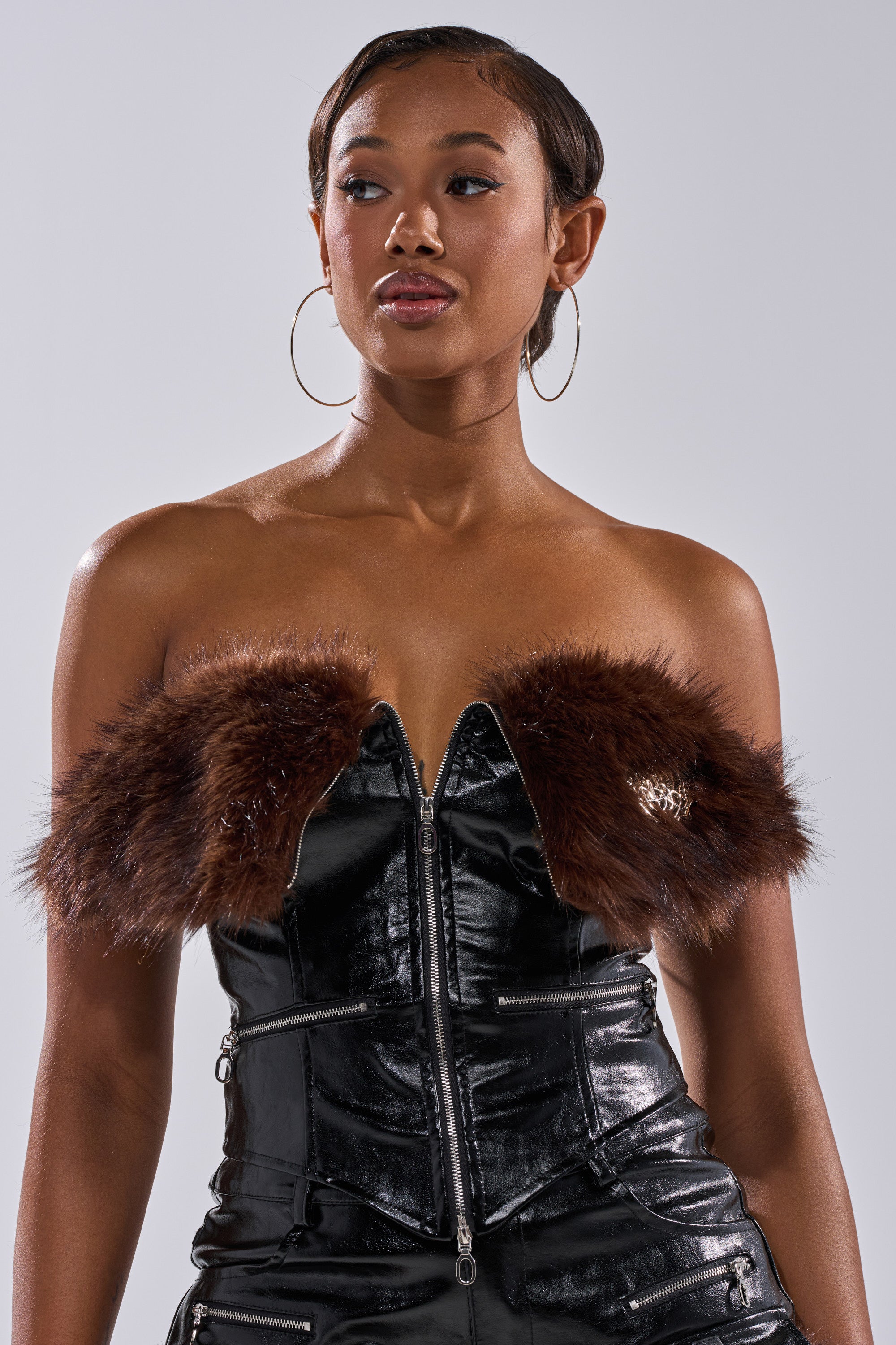 A woman poses against a plain background wearing the ALL THE BIG ONES CORSET WITH FUR—a strapless faux leather corset with a zip front, brown fur trim, and large hoop earrings.