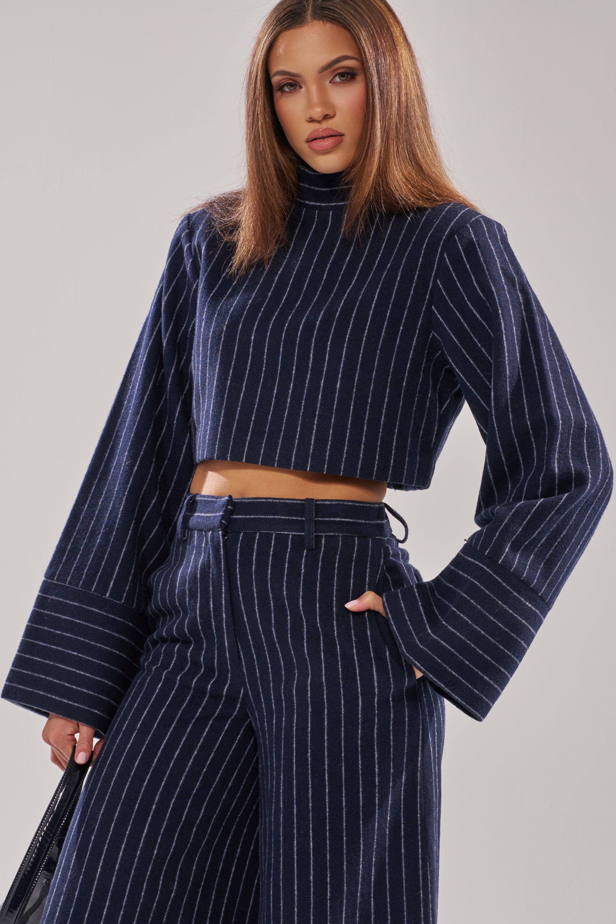 A woman with straight brown hair wears the STRICTLY BUSINESS PINSTRIPE BLOUSE, a navy blue, cropped pinstripe top paired with matching high-waisted wide-leg pants. She confidently holds a black bag against a plain background. 0