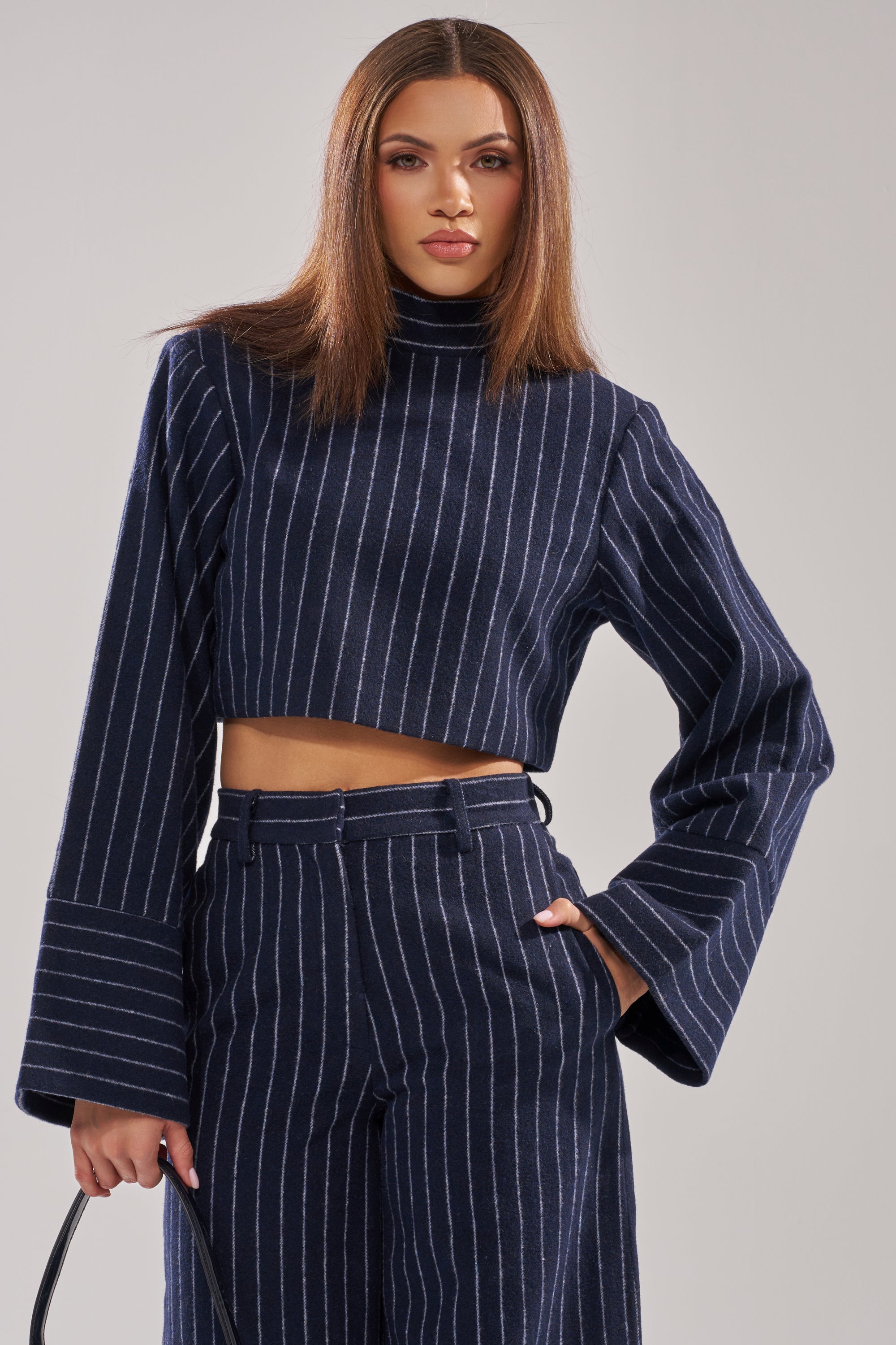 A woman stands confidently, holding a black handbag by her side, dressed in the STRICTLY BUSINESS PINSTRIPE BLOUSE—navy blue with wide sleeves and a cropped fit—paired with matching high-waisted pants. 1