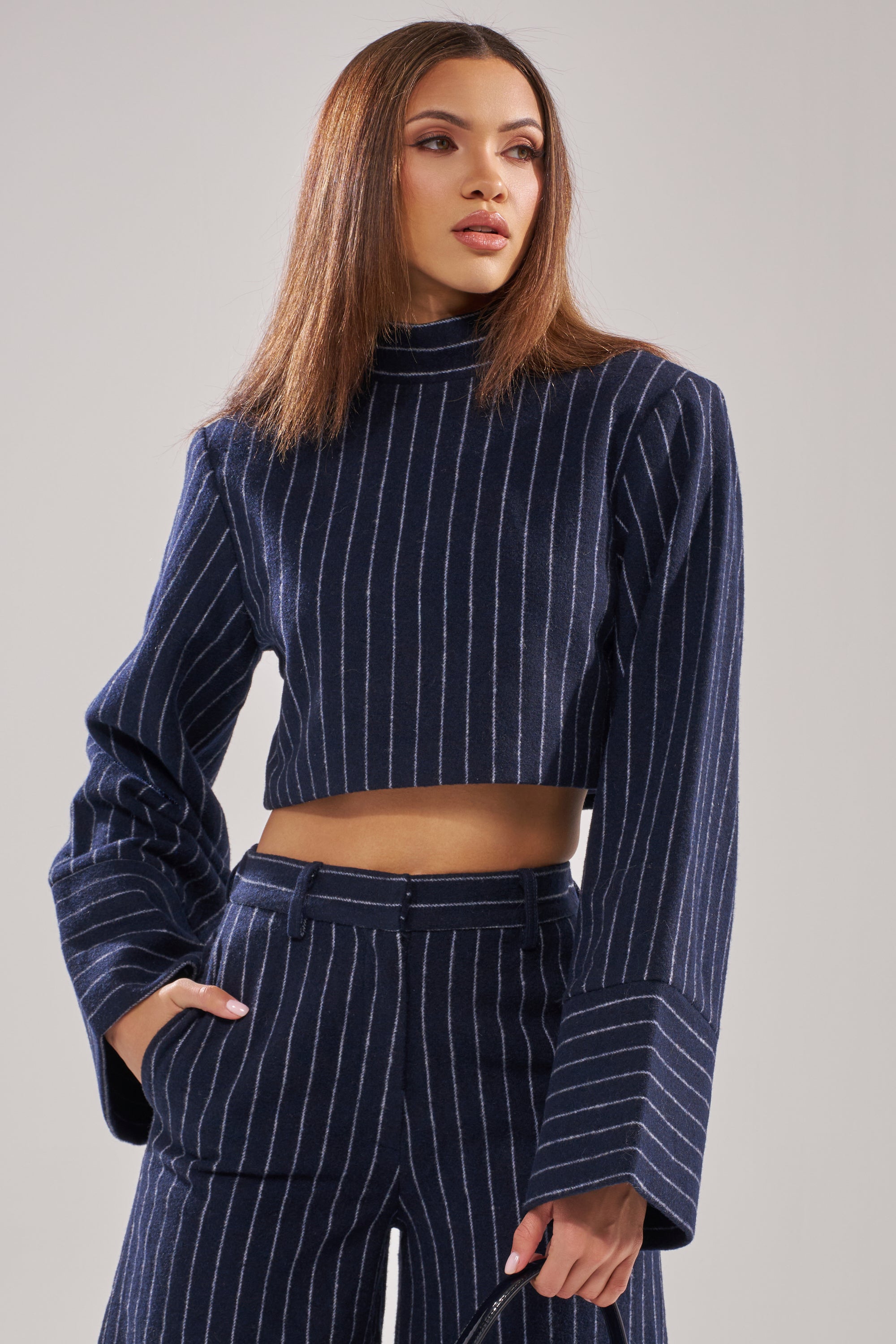A woman wearing the STRICTLY BUSINESS PINSTRIPE BLOUSE, a navy blue long-sleeve cropped top with matching high-waisted pants, stands confidently with one hand in her pocket against a plain background. 2