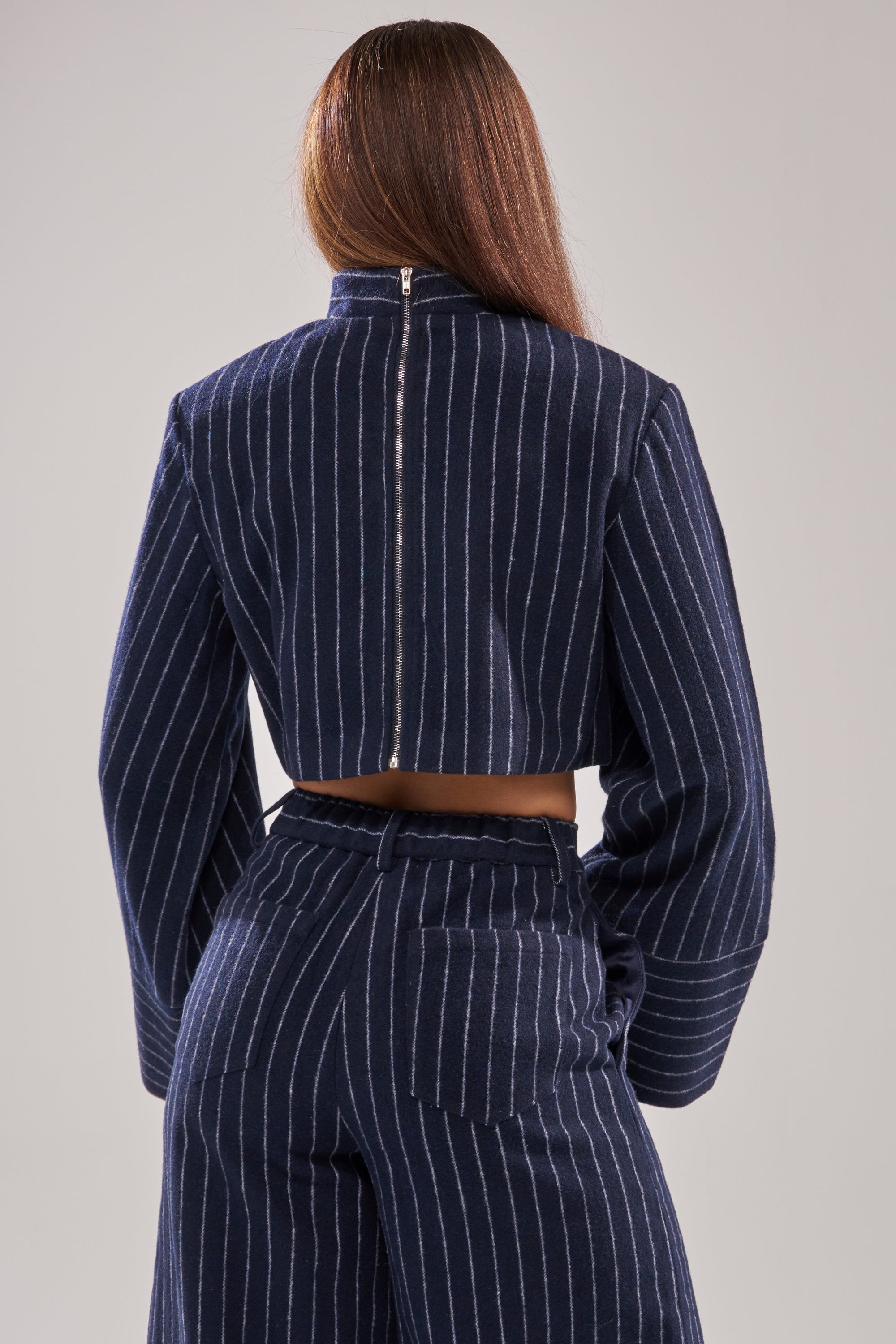 A person with long brown hair is seen from the back wearing the STRICTLY BUSINESS PINSTRIPE BLOUSE, a navy blue pinstripe cropped jacket with a back zipper, paired with high-waisted pants featuring rear pockets. 4