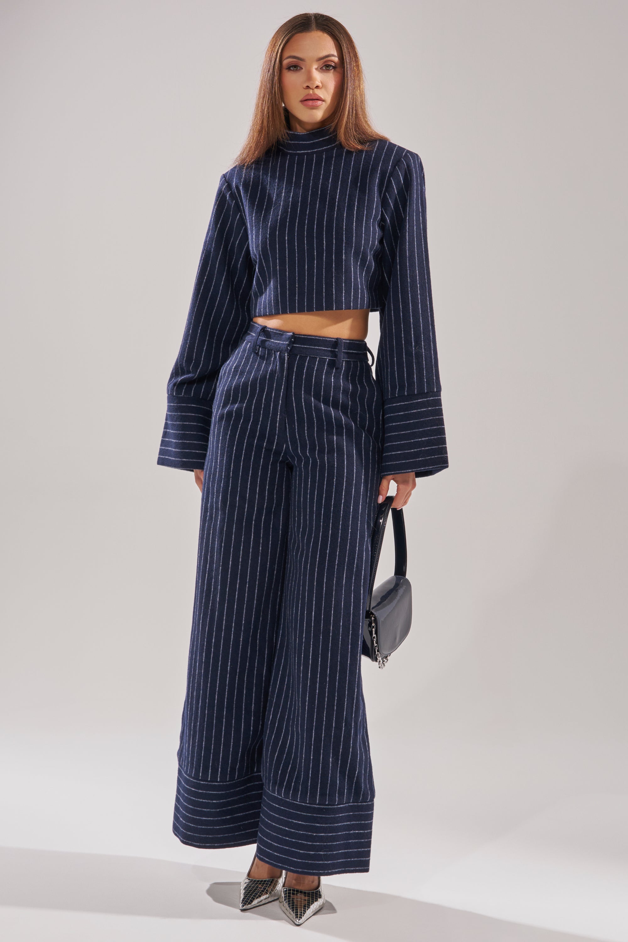 A woman models the STRICTLY BUSINESS PINSTRIPE BLOUSE in navy blue, paired with cropped wide-leg pants. She accessorizes with a small black handbag and pointed-toe heels. 5
