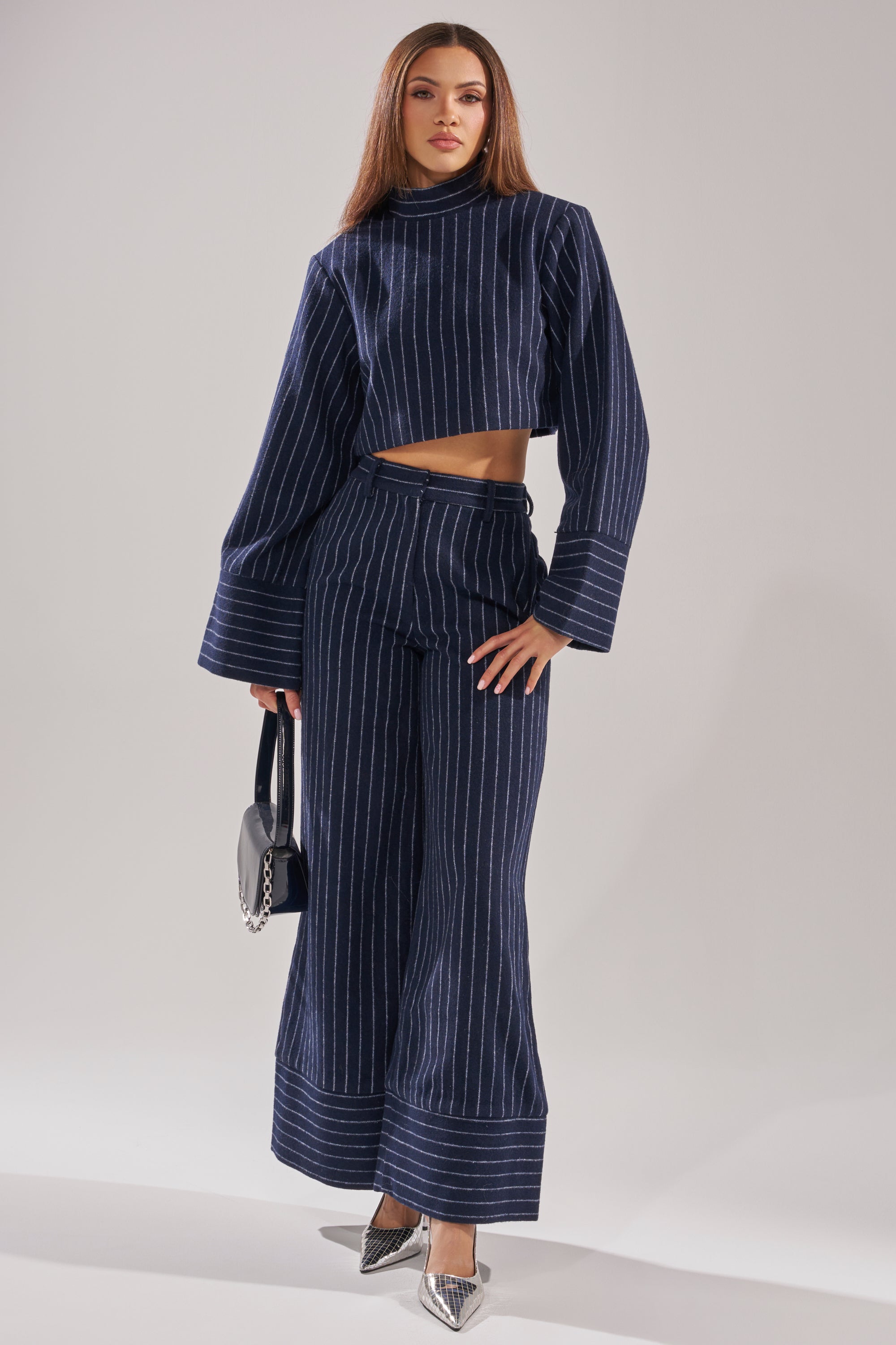 A woman wears the STRICTLY BUSINESS PINSTRIPE BLOUSE set—navy blue with pinstripes, featuring a cropped turtleneck top and wide-leg pants. She carries a small black handbag, pairs pointed silver heels, and stands against a plain light background. 6
