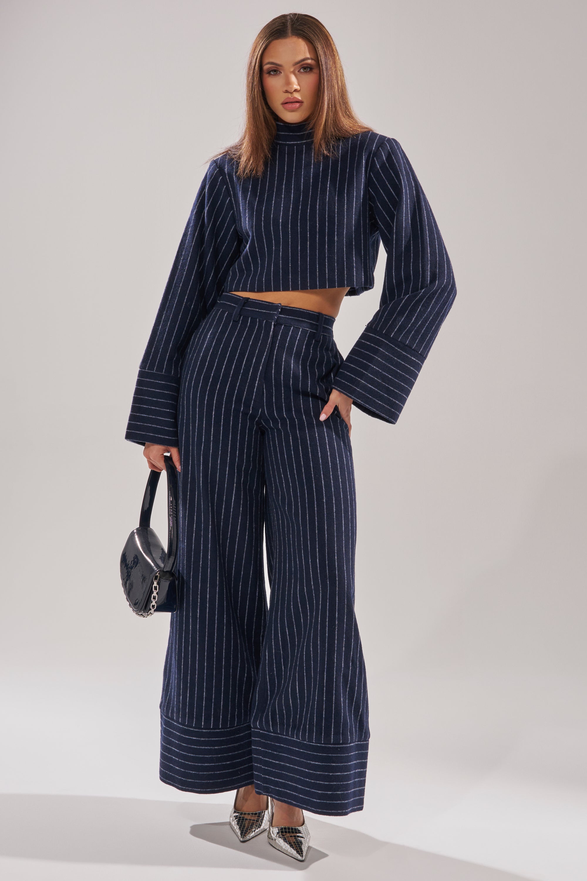 A woman models the STRICTLY BUSINESS PINSTRIPE BLOUSE with a cropped fit, paired with wide-leg pants. She accessorizes with a small black handbag and metallic pointed-toe heels against a plain, light-colored background. 7