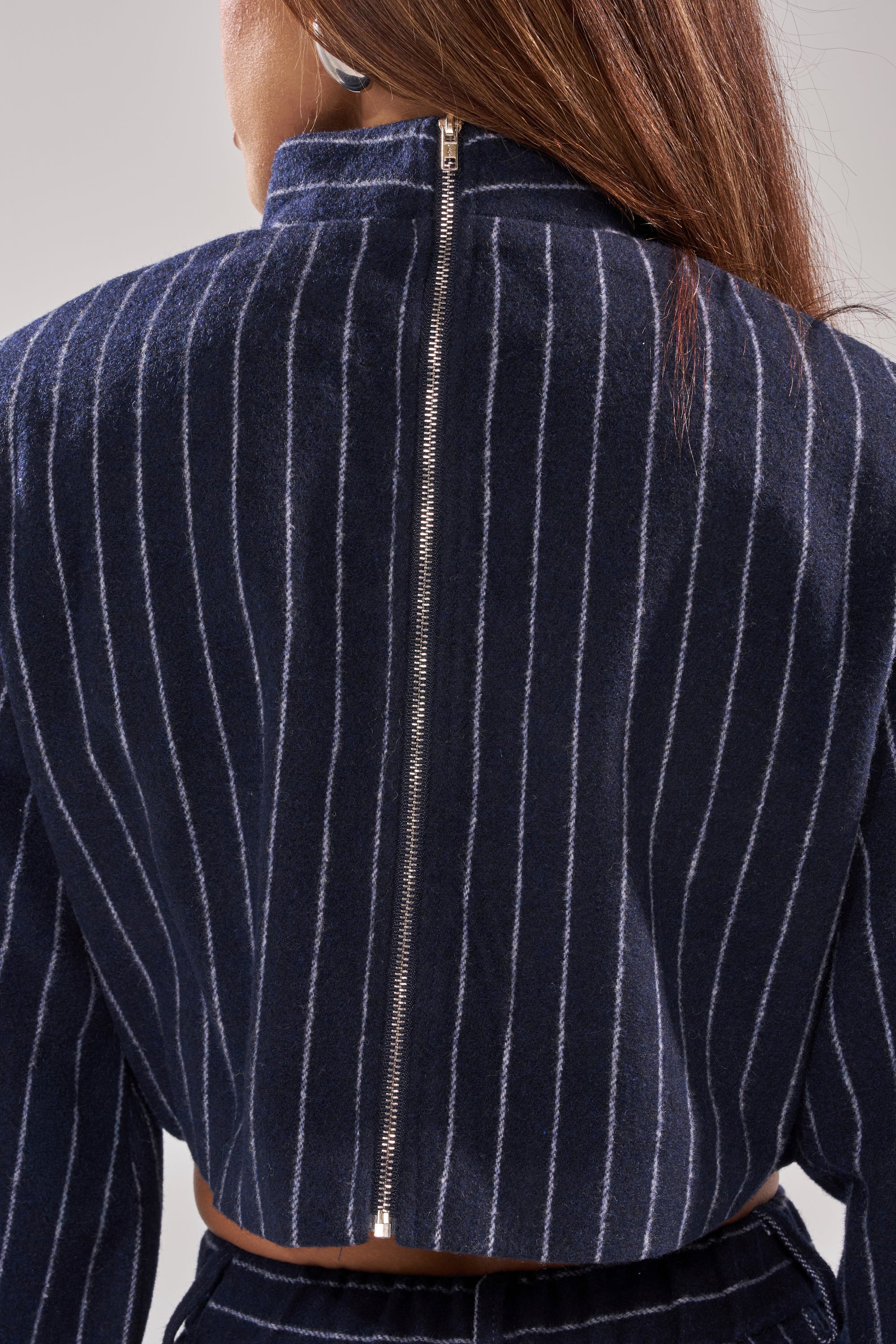 A person wears the STRICTLY BUSINESS PINSTRIPE BLOUSE, featuring dark blue pinstripes and a visible silver back zipper. The image highlights the textured fabric, tailored shoulders, and chic cropped fit. 8