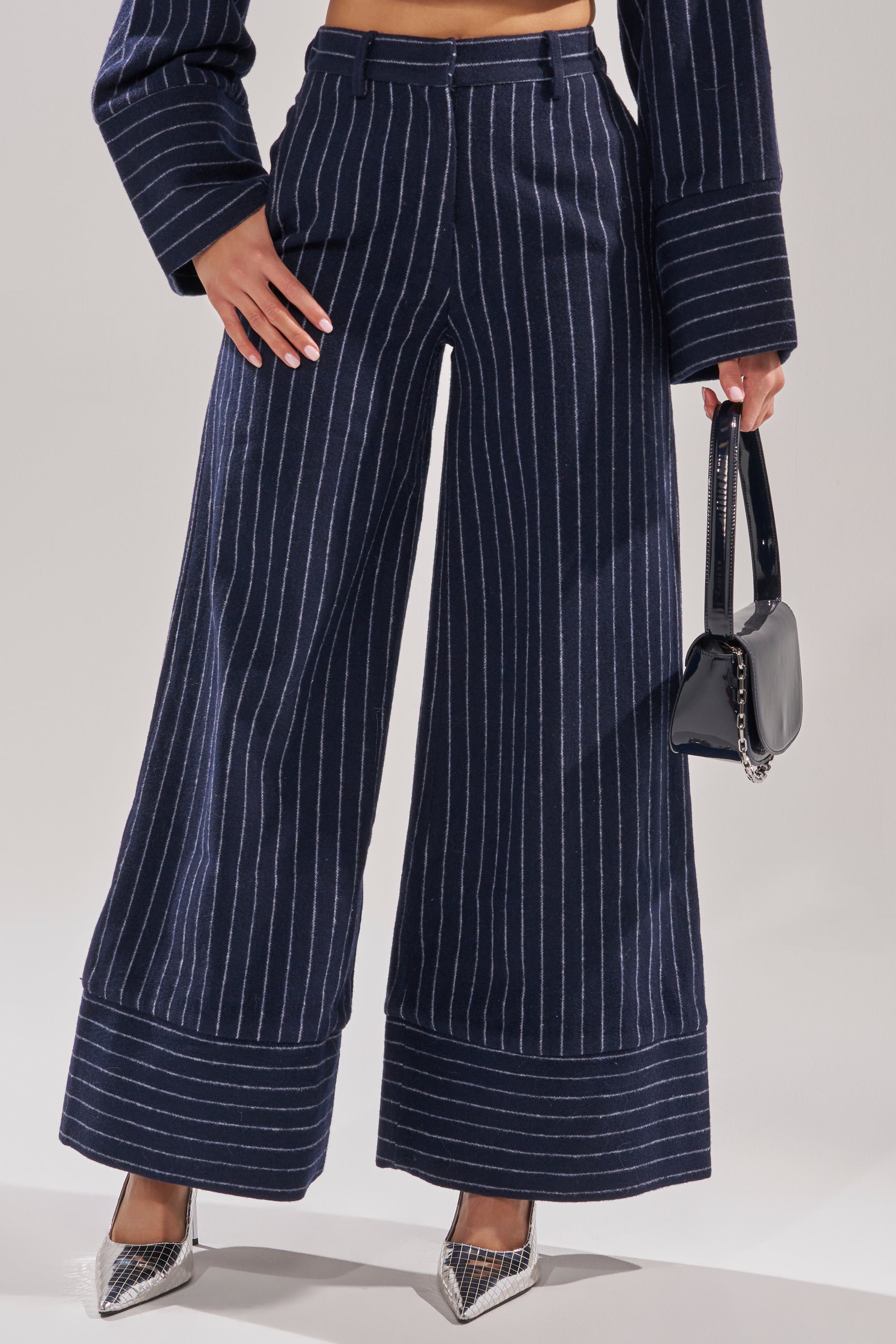 Wearing the STRICTLY BUSINESS PINSTRIPE TROUSER and matching jacket, a person holds a small black handbag and sports pointed silver heels. The photo is cropped at the shoulders. 1