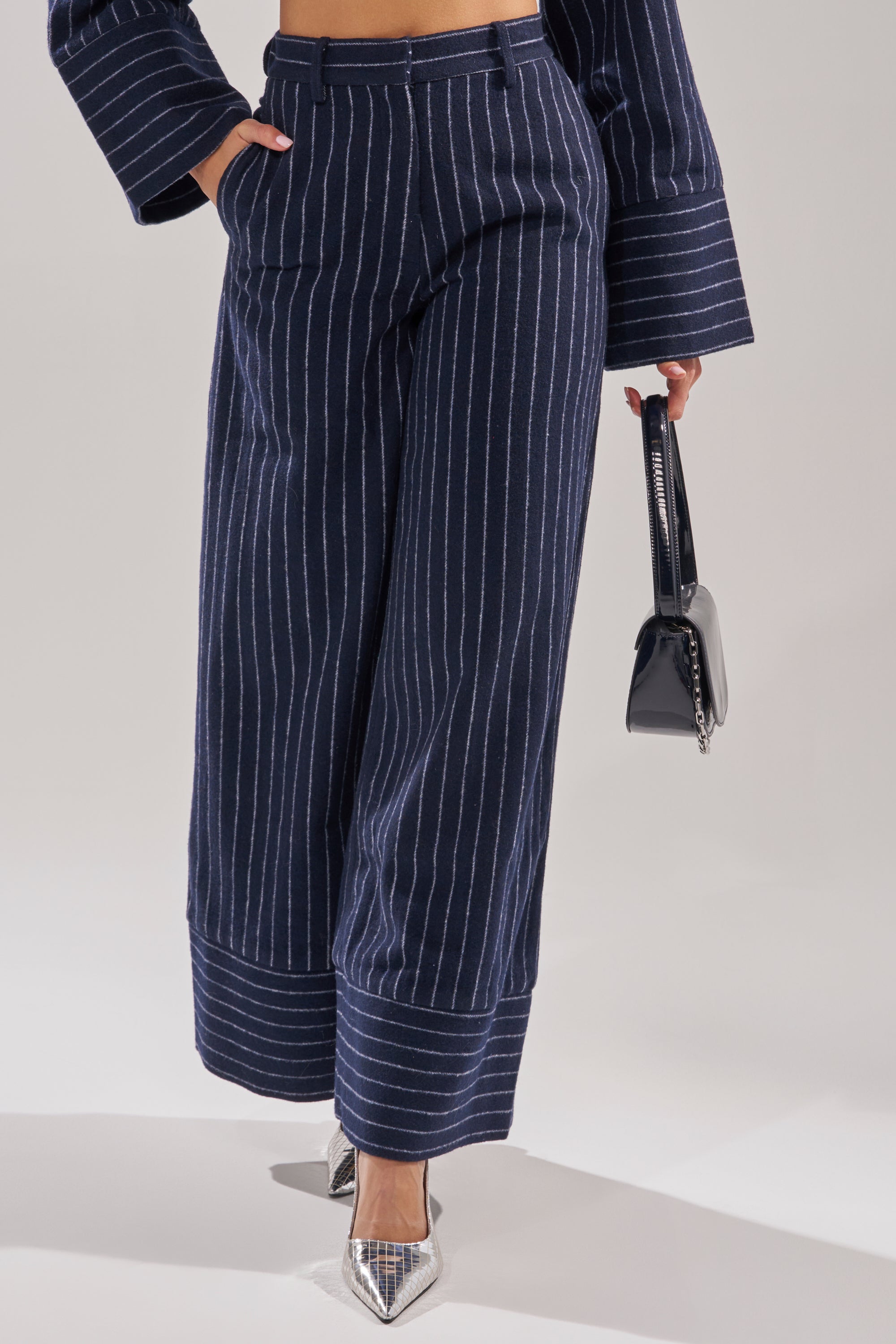 A person wears the STRICTLY BUSINESS PINSTRIPE TROUSER in navy blue with a high-rise fit, a matching long-sleeve top, silver pointed-toe heels, and holds a small black handbag. The photo is cropped from shoulders to feet. 2