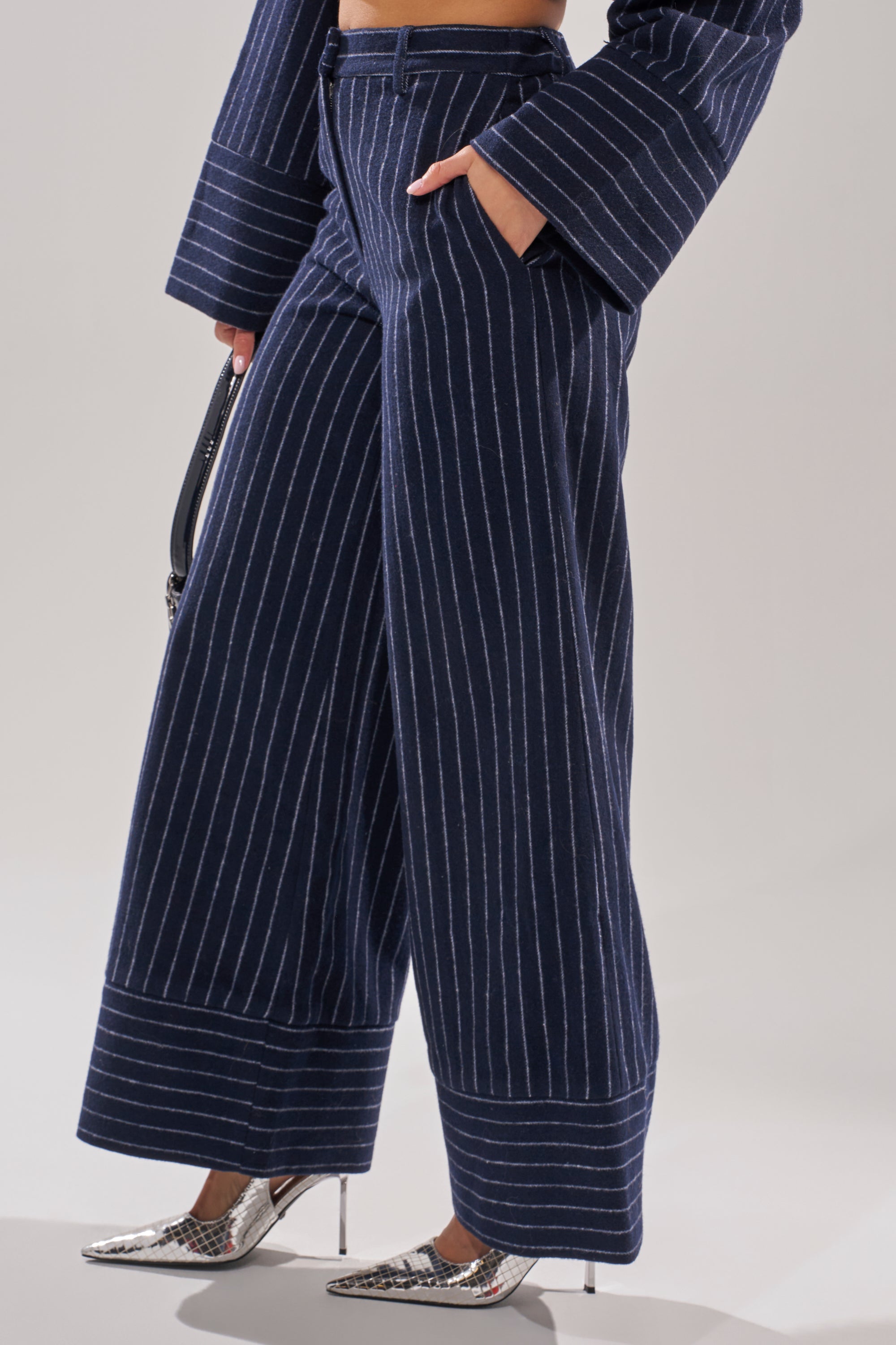 Someone wears the STRICTLY BUSINESS PINSTRIPE TROUSER in navy blue with a matching jacket, paired with a black handbag and silver pointed-toe heels, set against a plain light background. 3