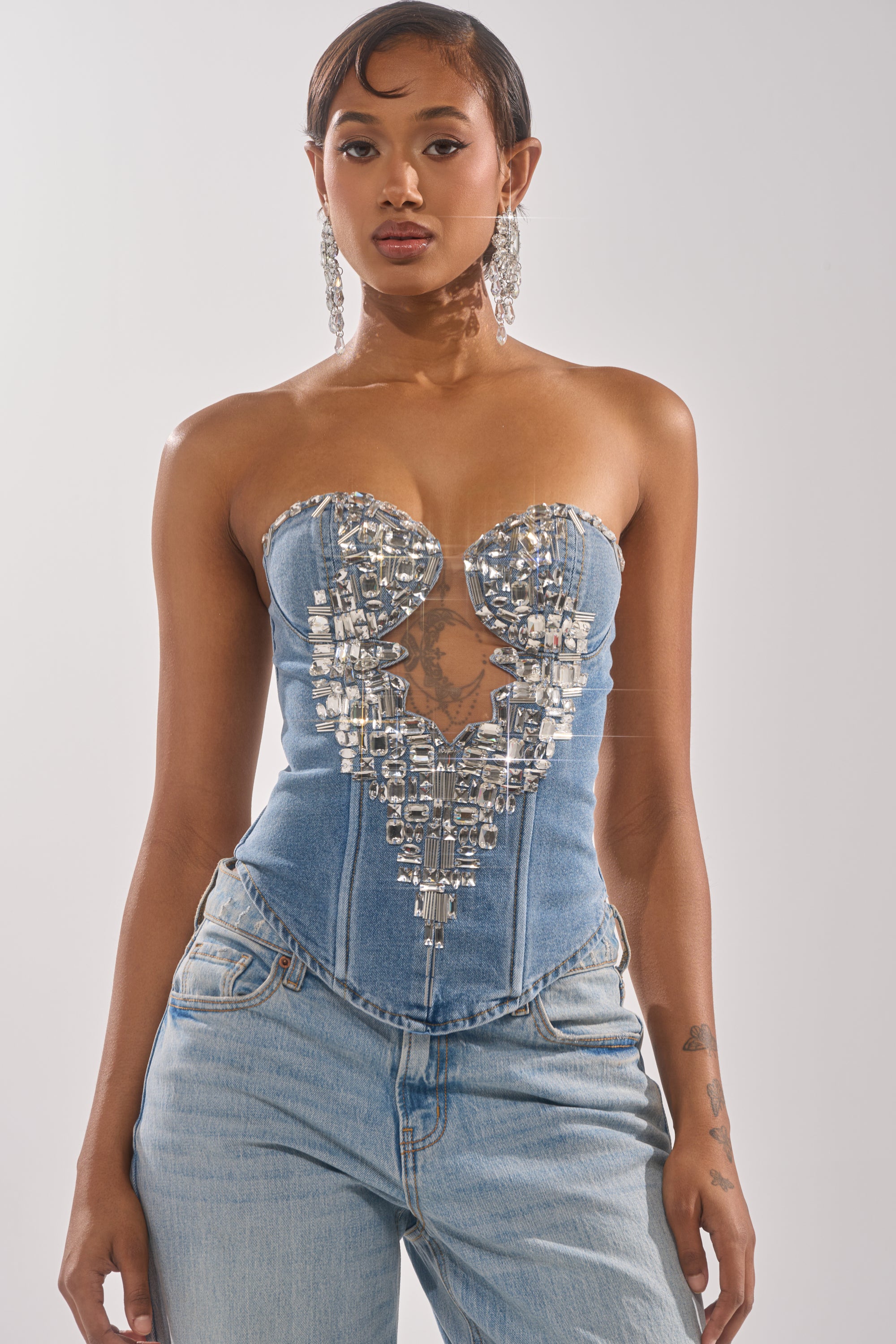 A woman stands facing forward in the GEMIMA CORSET, a strapless denim piece with a sweetheart neckline and silver embellishments, paired with light blue jeans. She has short styled hair and wears large sparkly earrings against a plain background.
