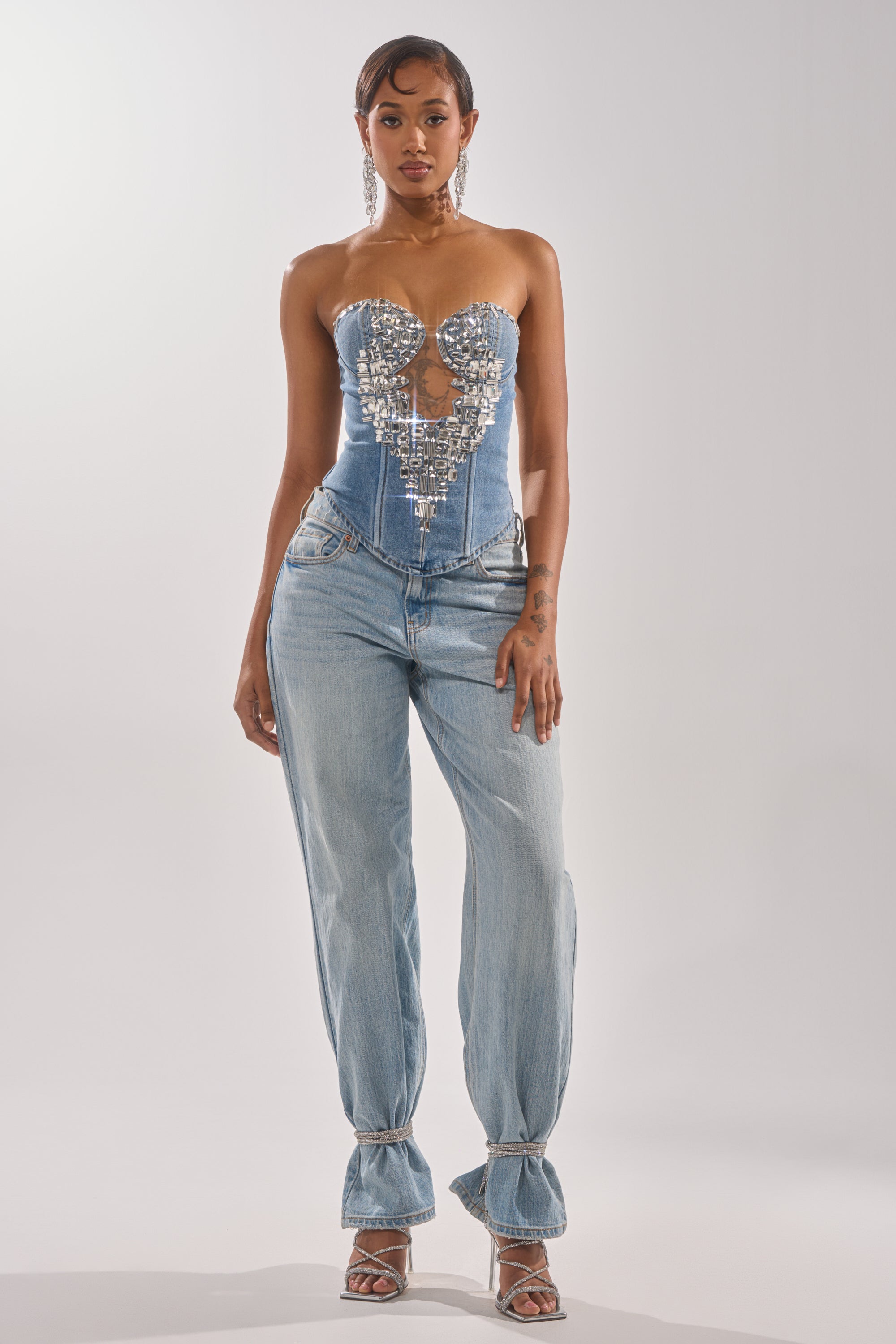 A woman faces forward in the GEMIMA CORSET, a strapless denim top with shiny silver embellishments, paired with light blue ankle-tied jeans. She wears hoop earrings, high heels, and has her hair pulled back against a plain light background.