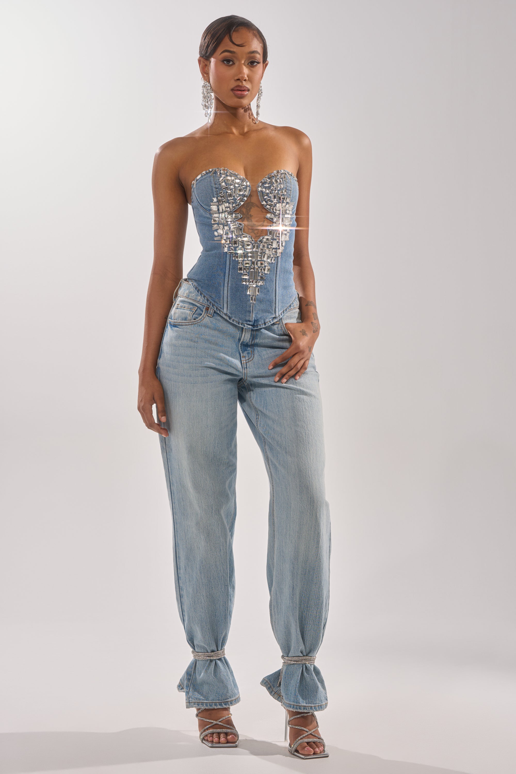 Wearing the GEMIMA CORSET, a strapless denim top with gemstone details, and light-wash loose jeans tied at the ankles, a woman with short hair poses in heeled sandals against a plain light background.