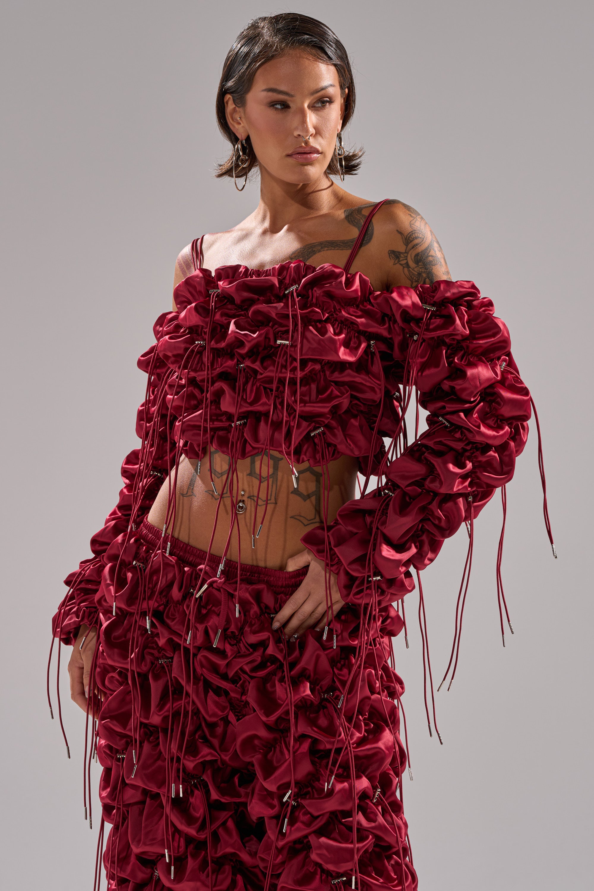 A person with short dark hair and tattoos models the FEELING FUNKY TIE BLOUSE, an off-the-shoulder, deep red ruched top with drawstrings, styled with a matching skirt, against a plain background.