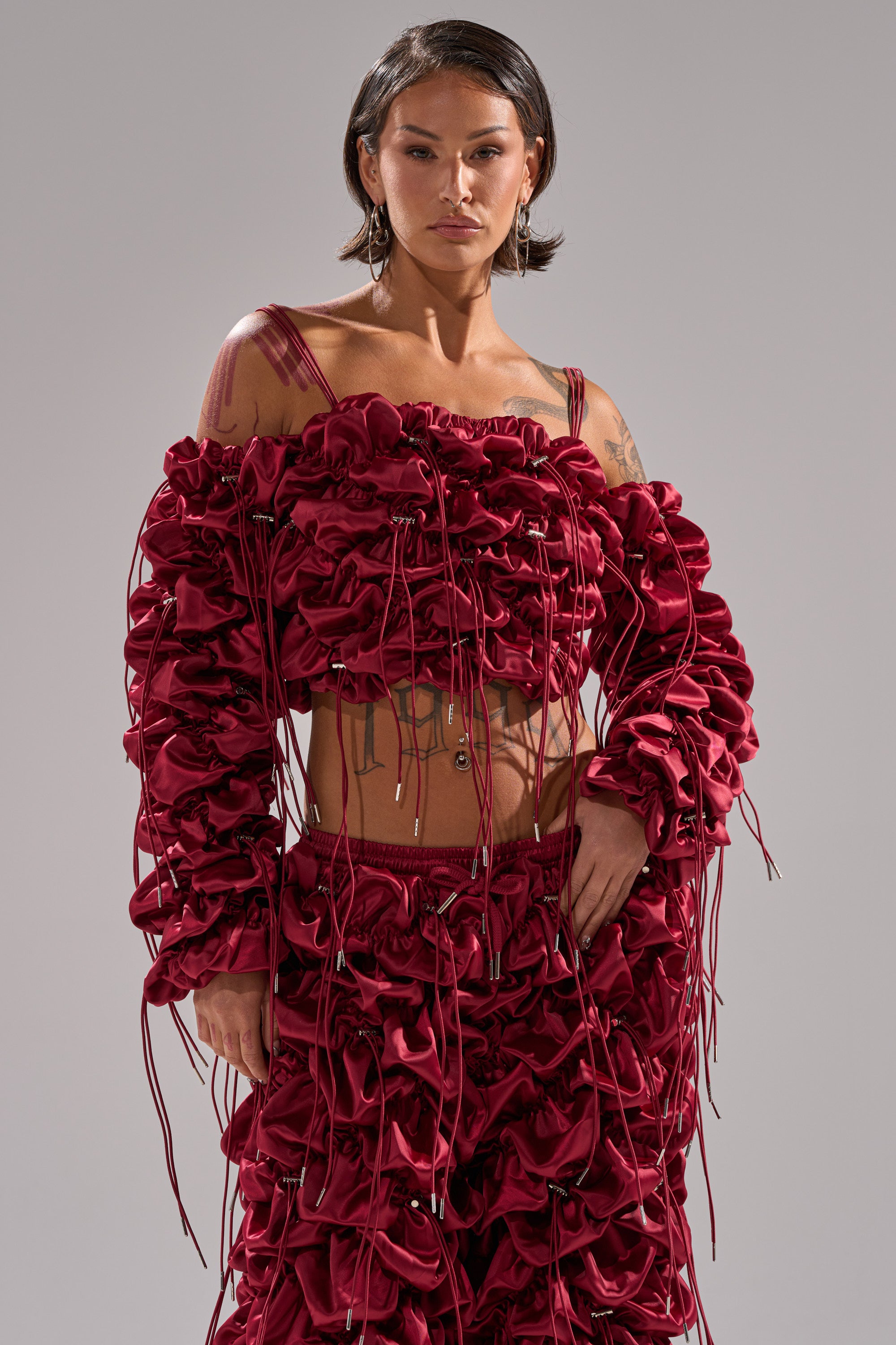 A person with short dark hair confidently poses in the FEELING FUNKY TIE BLOUSE, a bold deep red top with ruffled fabric and string sleeve details, paired with pants, against a plain light gray background.