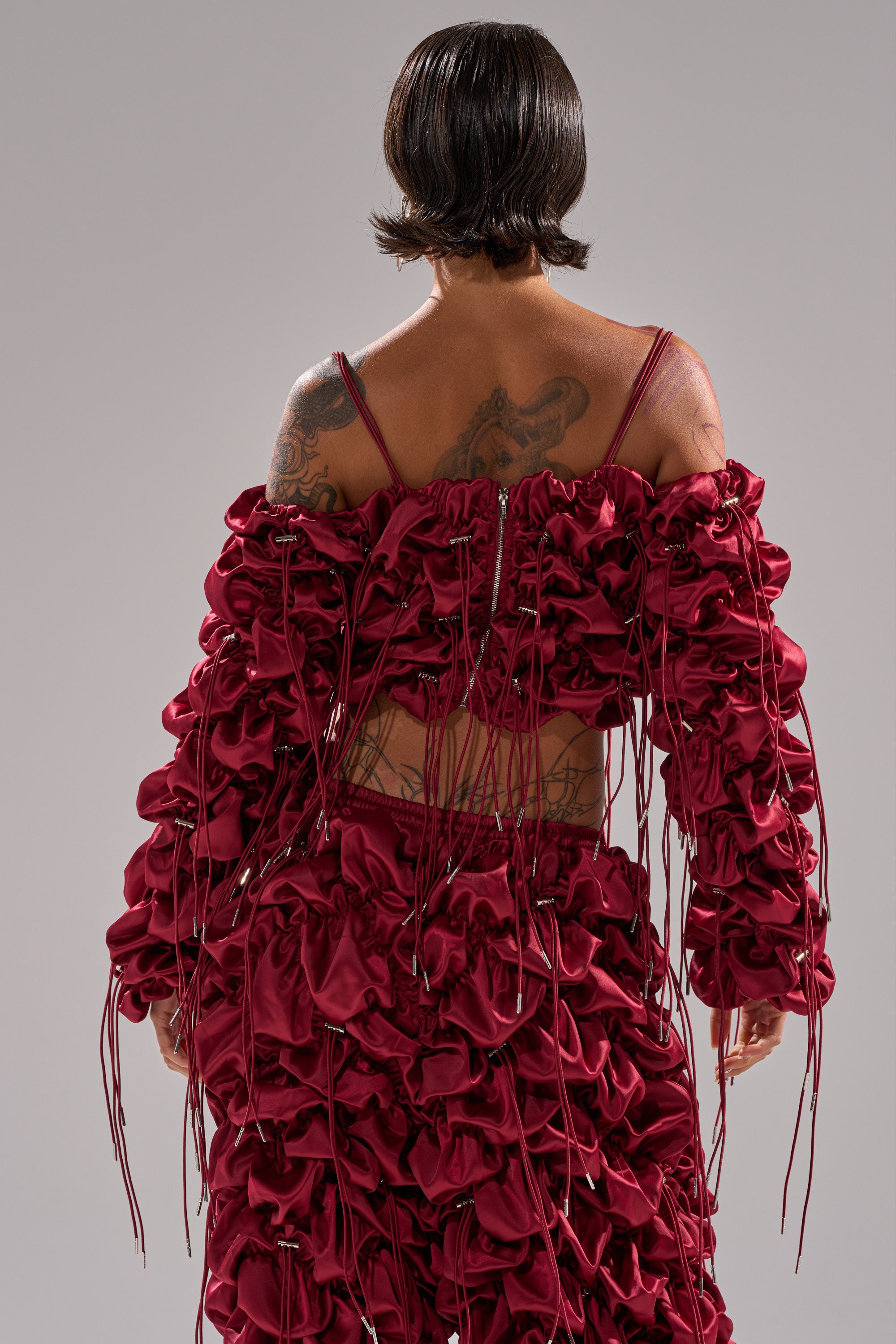 A person with short, dark hair and tattoos is seen from behind, wearing the FEELING FUNKY TIE BLOUSE in deep red ruched satin with multiple strings, set against a plain gray background.