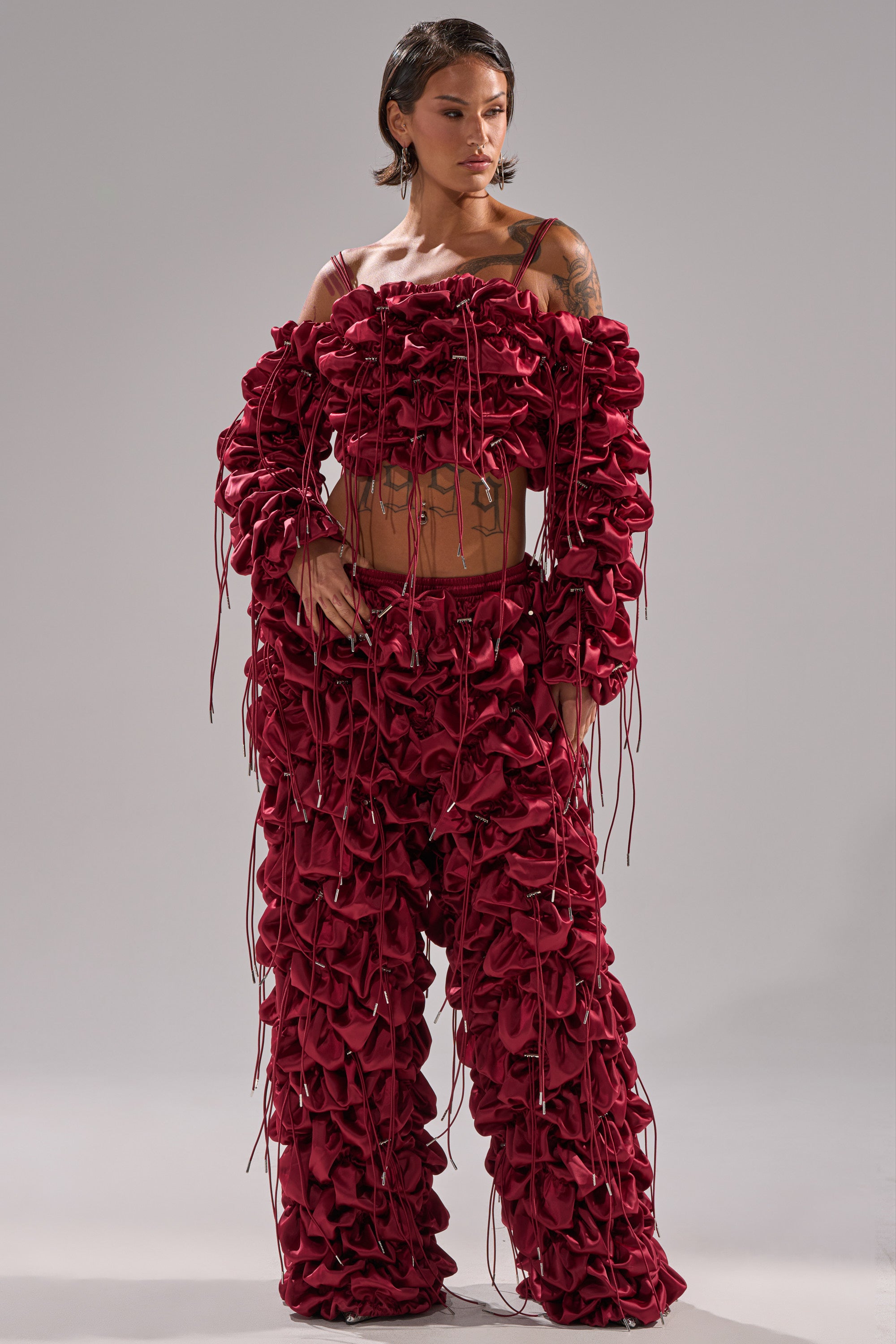 A person stands in a studio against a plain gray background wearing the FEELING FUNKY TIE BLOUSE, a bold deep red ruched satin top with gathered ruffles, long dangling strings, and arm coverage while revealing the midriff.