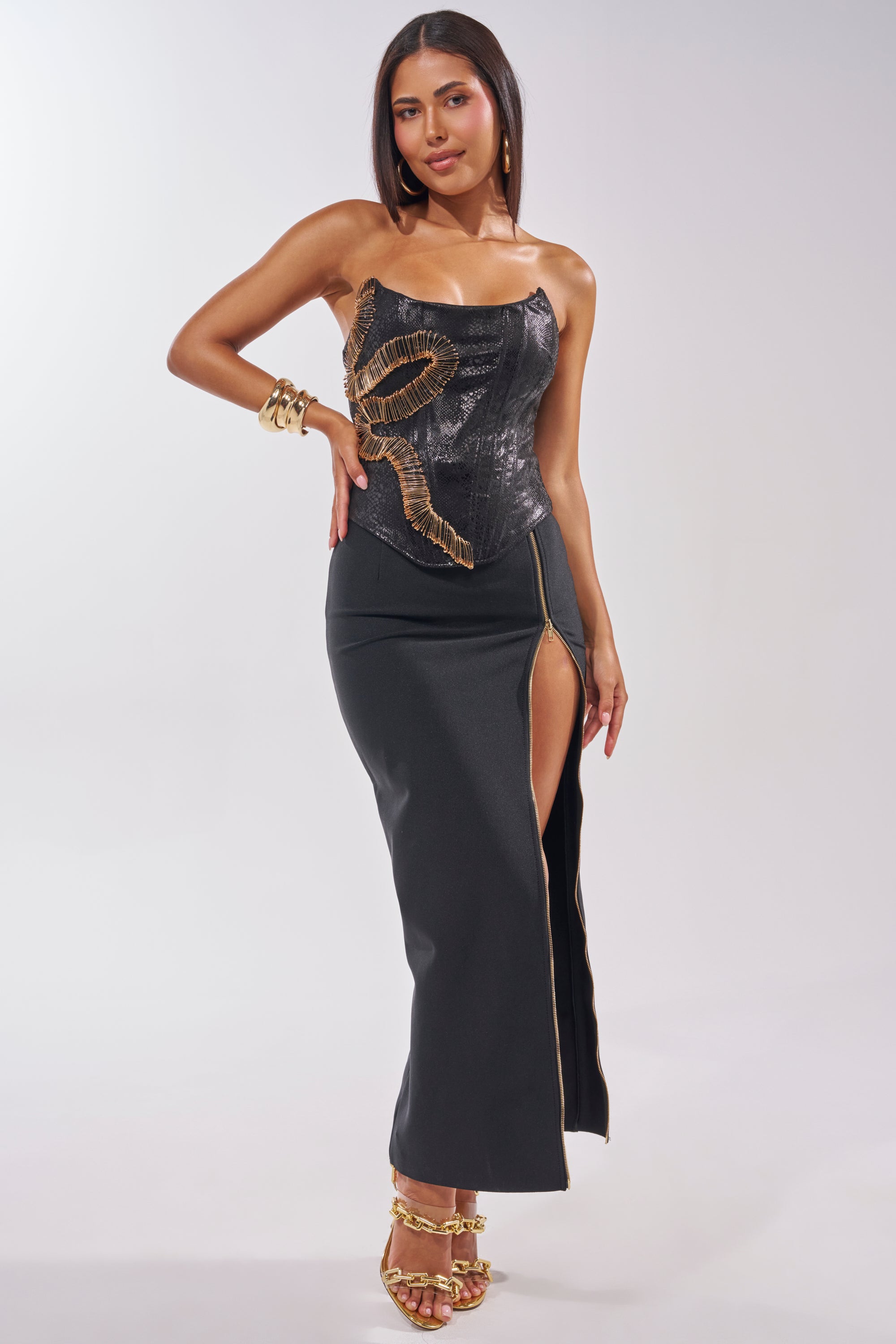 A woman poses in the VENOMOUS EMBELLISHED PU CORSET, featuring a gold snake design, high front slit, styled with gold sandals, hoop earrings, and bracelets against a plain light background. 6