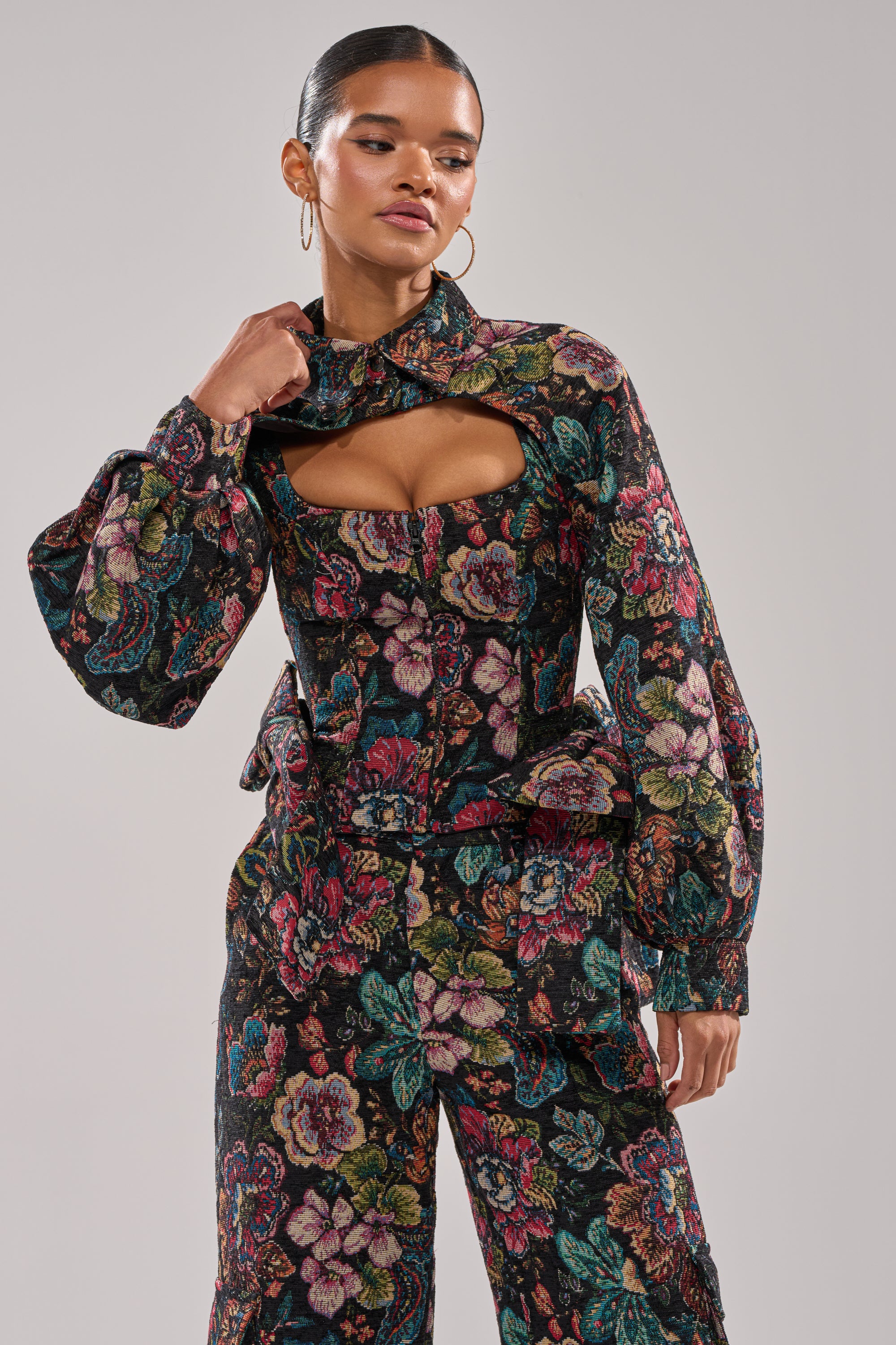 A woman models the PRETTY LADY BROCADE BOLERO with puffed sleeves and matching wide-leg floral pants, showcasing a unique chest cutout, posing confidently against a simple backdrop. 0