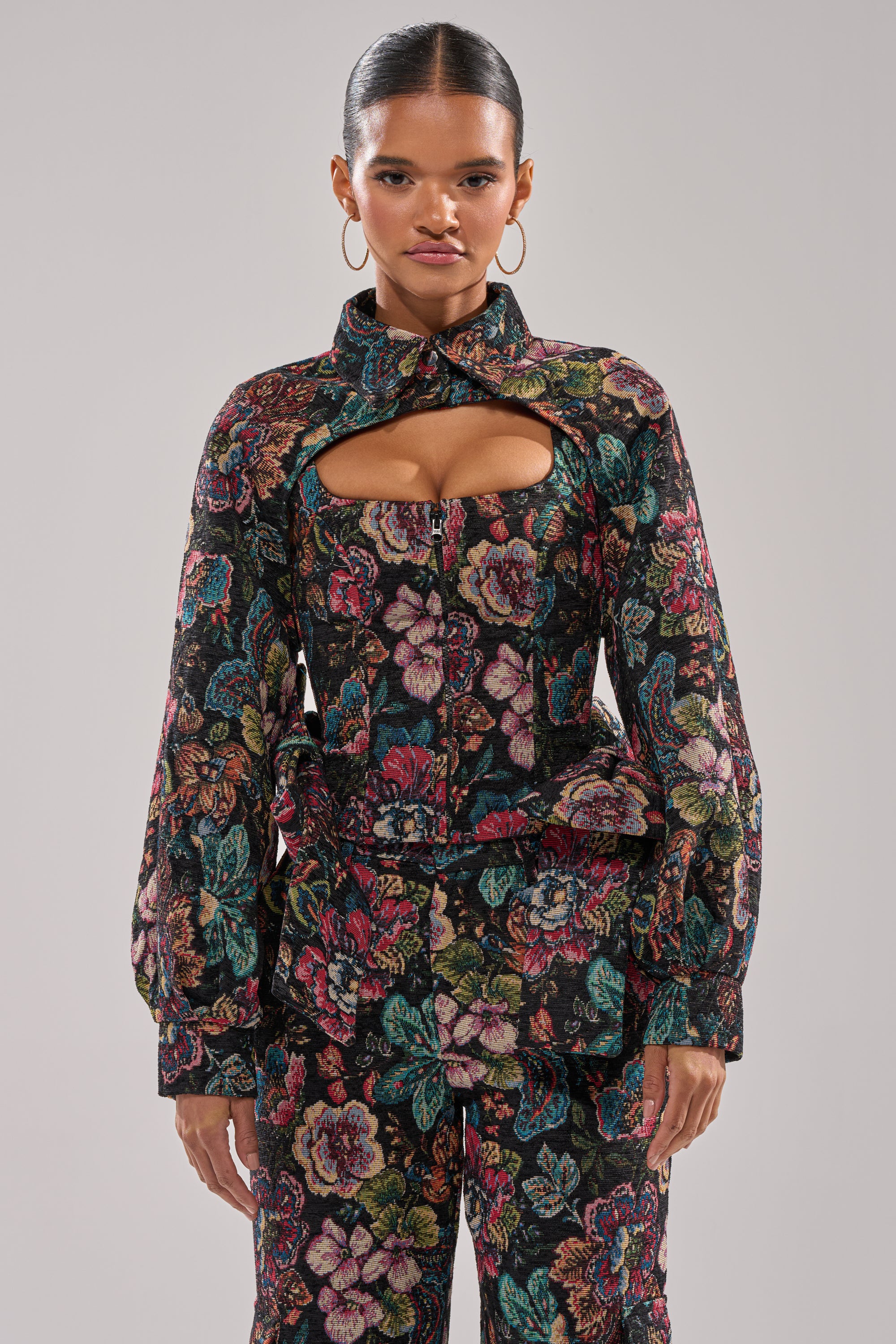 A woman faces forward in the PRETTY LADY BROCADE BOLERO, featuring a floral pattern, high collar, chest cut-out, long sleeves, matching pants, and hoop earrings. Her hair is slicked back for a chic look. 1