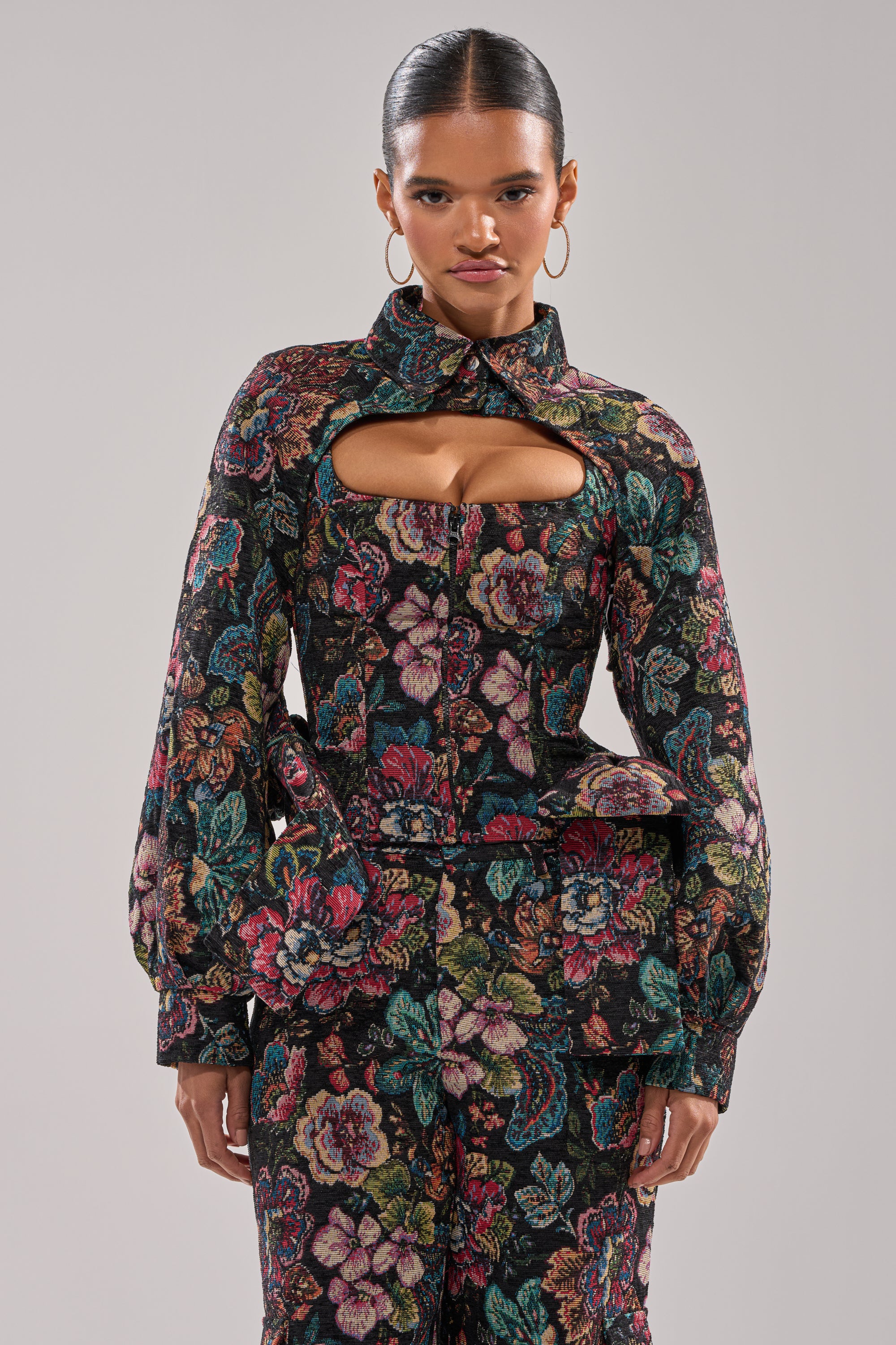A person with slicked-back hair wears the PRETTY LADY BROCADE BOLERO— a black, high-neck floral jacket with chest cut-out and long puffed sleeves—paired with matching pants. They stand confidently against a neutral background. 2