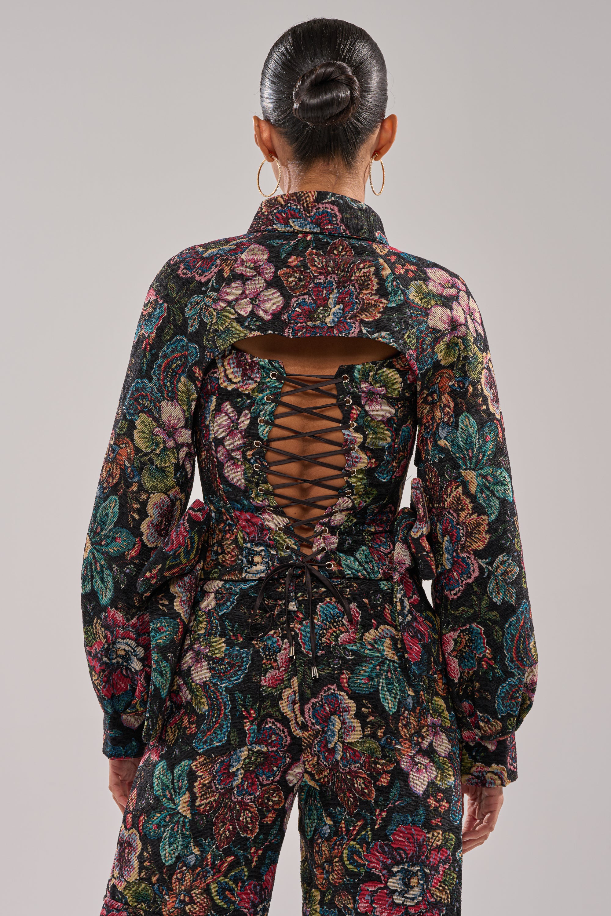 A person stands facing away, wearing the PRETTY LADY BROCADE BOLERO with long sleeves and floral pattern. The back features a large cutout and crisscross lace-up detail. Their hair is in a sleek, low bun. 4