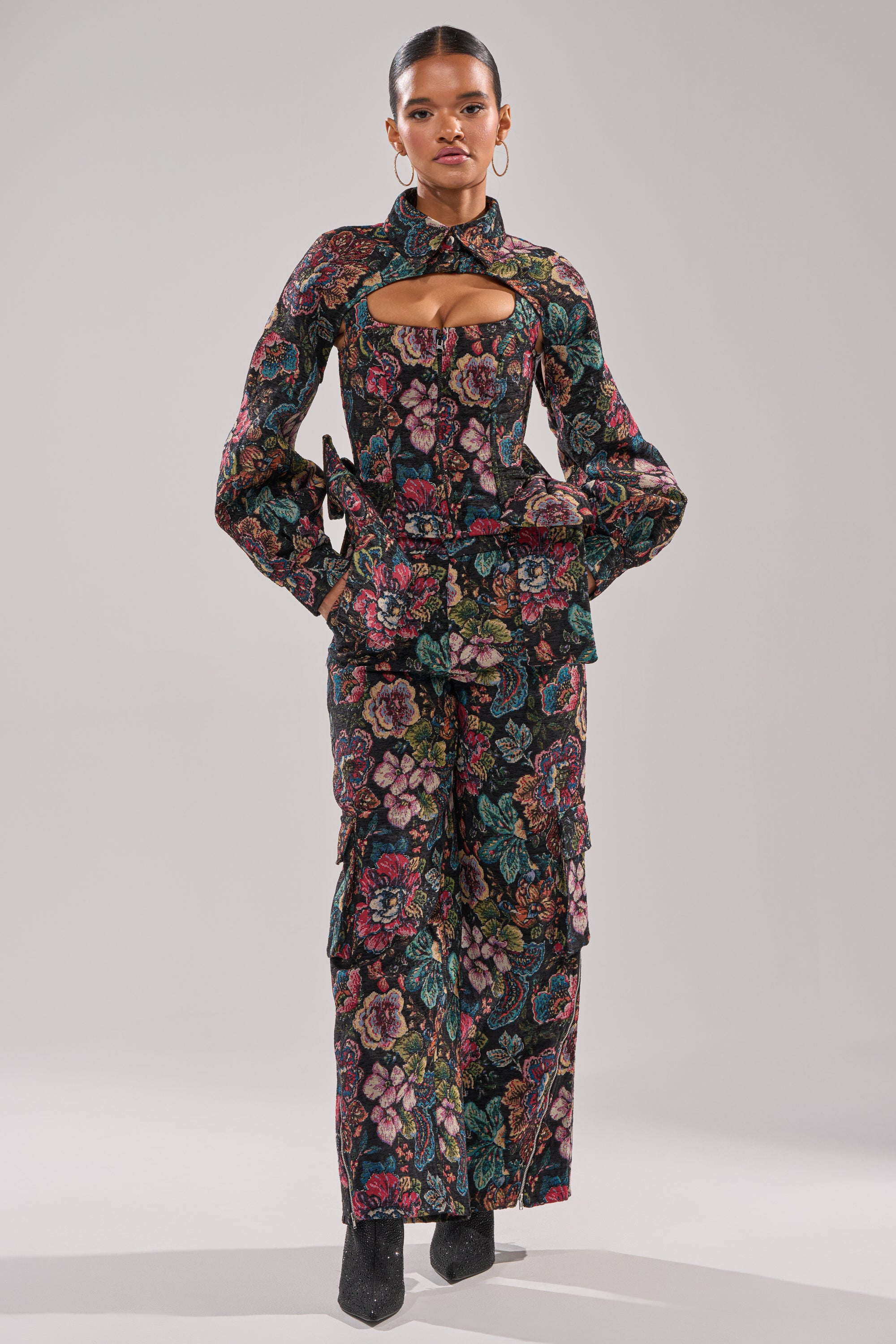 A woman poses against a plain light background wearing the PRETTY LADY BROCADE BOLERO as part of a matching floral pantsuit with a chest cutout, long sleeves, wide-leg pants, and black pointed-toe boots. 6