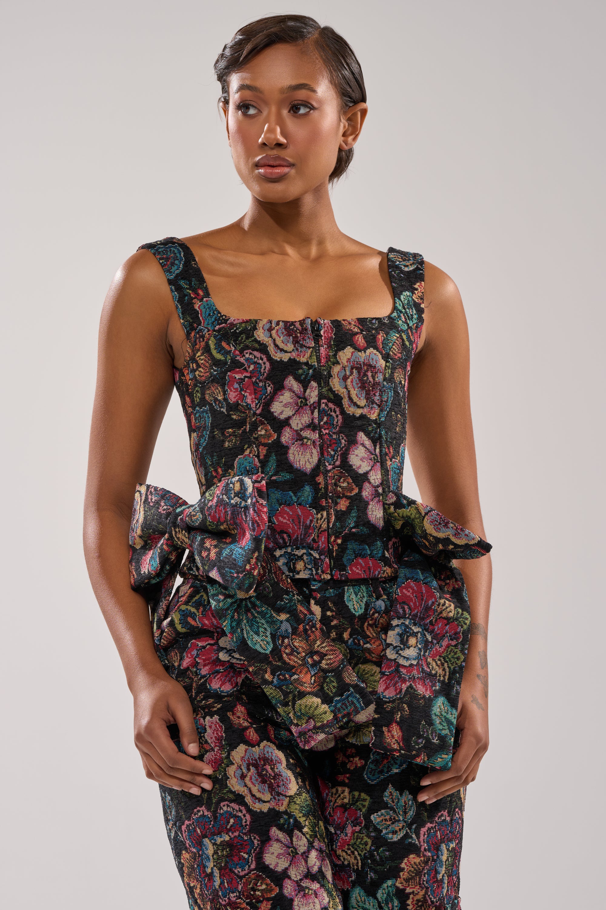 A woman poses against a plain light background wearing the PRETTY LITTLE LADY BROCADE CORSET, a floral cropped top with a square neckline, styled with matching pants featuring a large decorative waist ruffle. 0