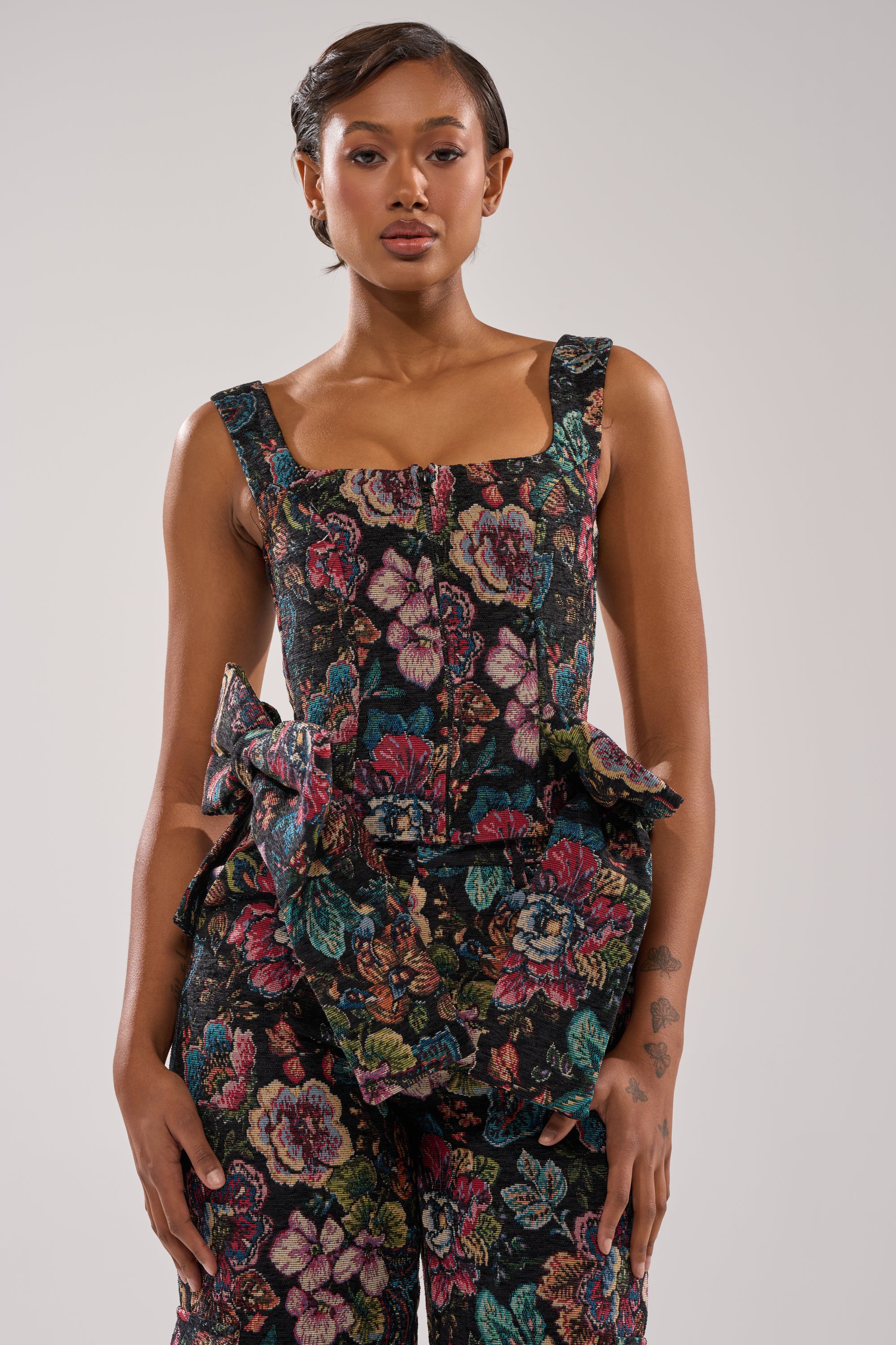 A woman in the PRETTY LITTLE LADY BROCADE CORSET, featuring a sleeveless, square neckline and colorful floral pattern with ruffle waist details, stands against a plain light background, confidently looking at the camera. 1