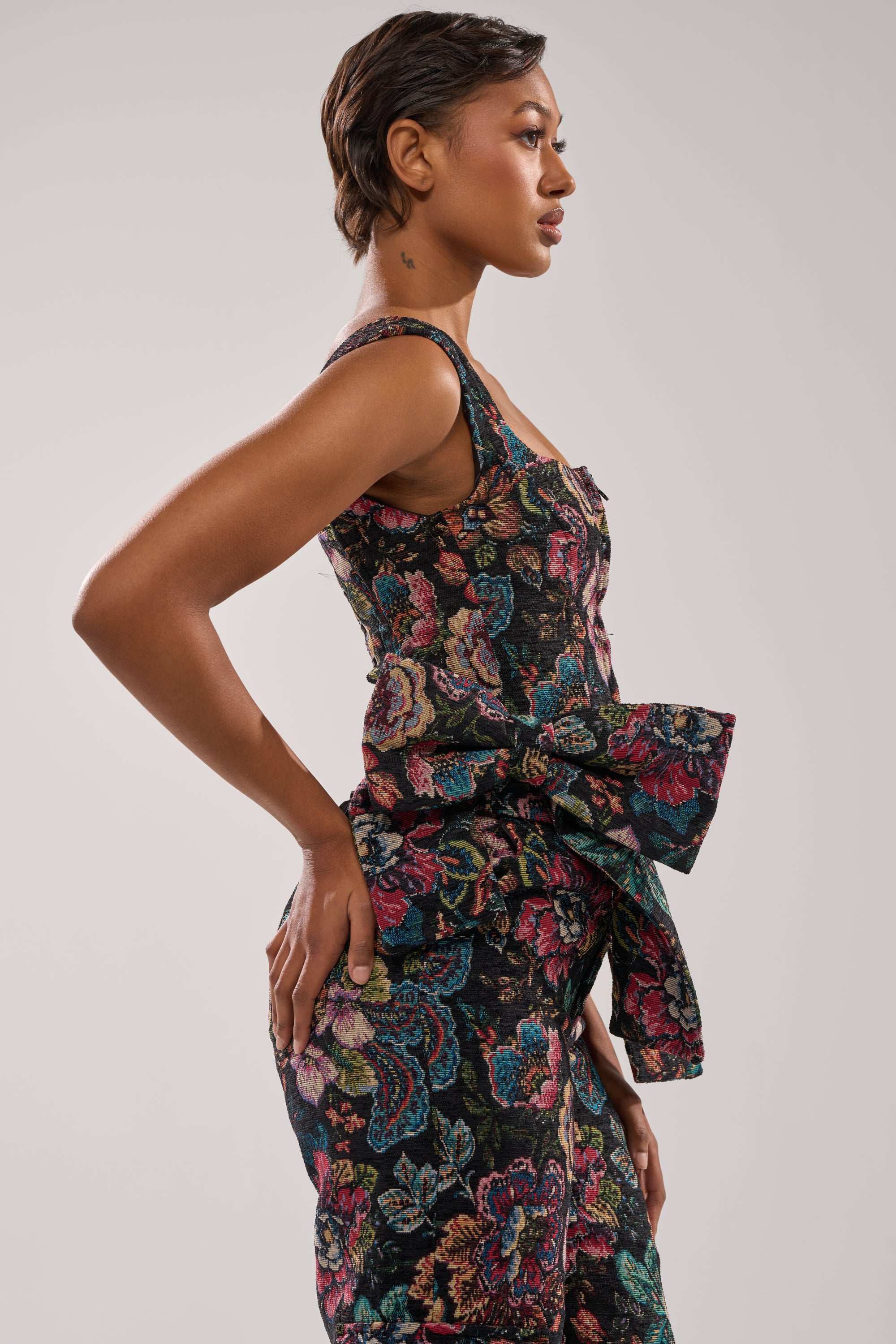 A woman in profile showcases the PRETTY LITTLE LADY BROCADE CORSET, a sleeveless, fitted jumpsuit featuring a colorful floral design and a large waist bow, standing against a light gray background. 2
