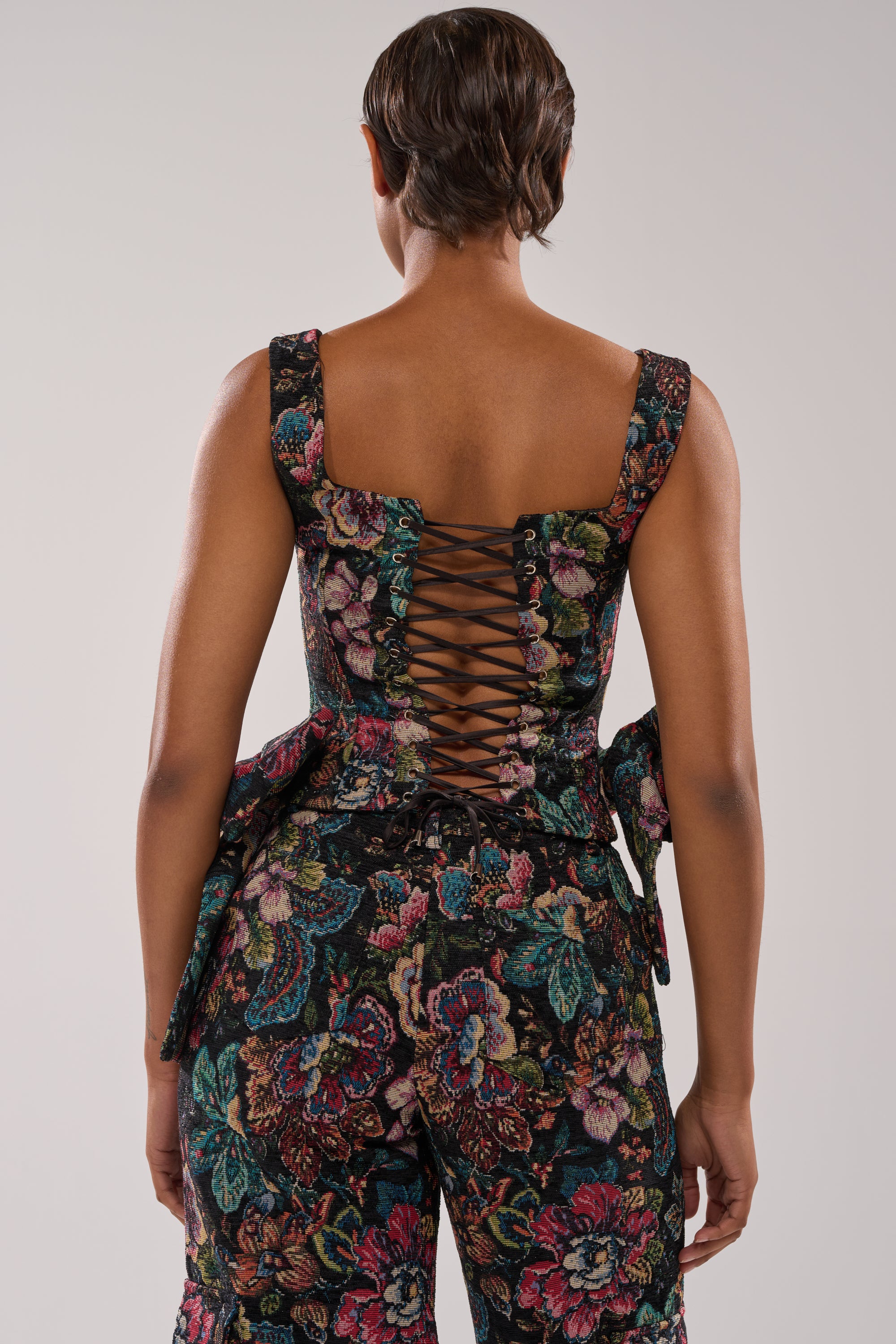 A person with short hair is shown from the back, wearing the PRETTY LITTLE LADY BROCADE CORSET, a sleeveless, floral corset-style top with a lace-up back and matching floral pants against a plain light background. 3