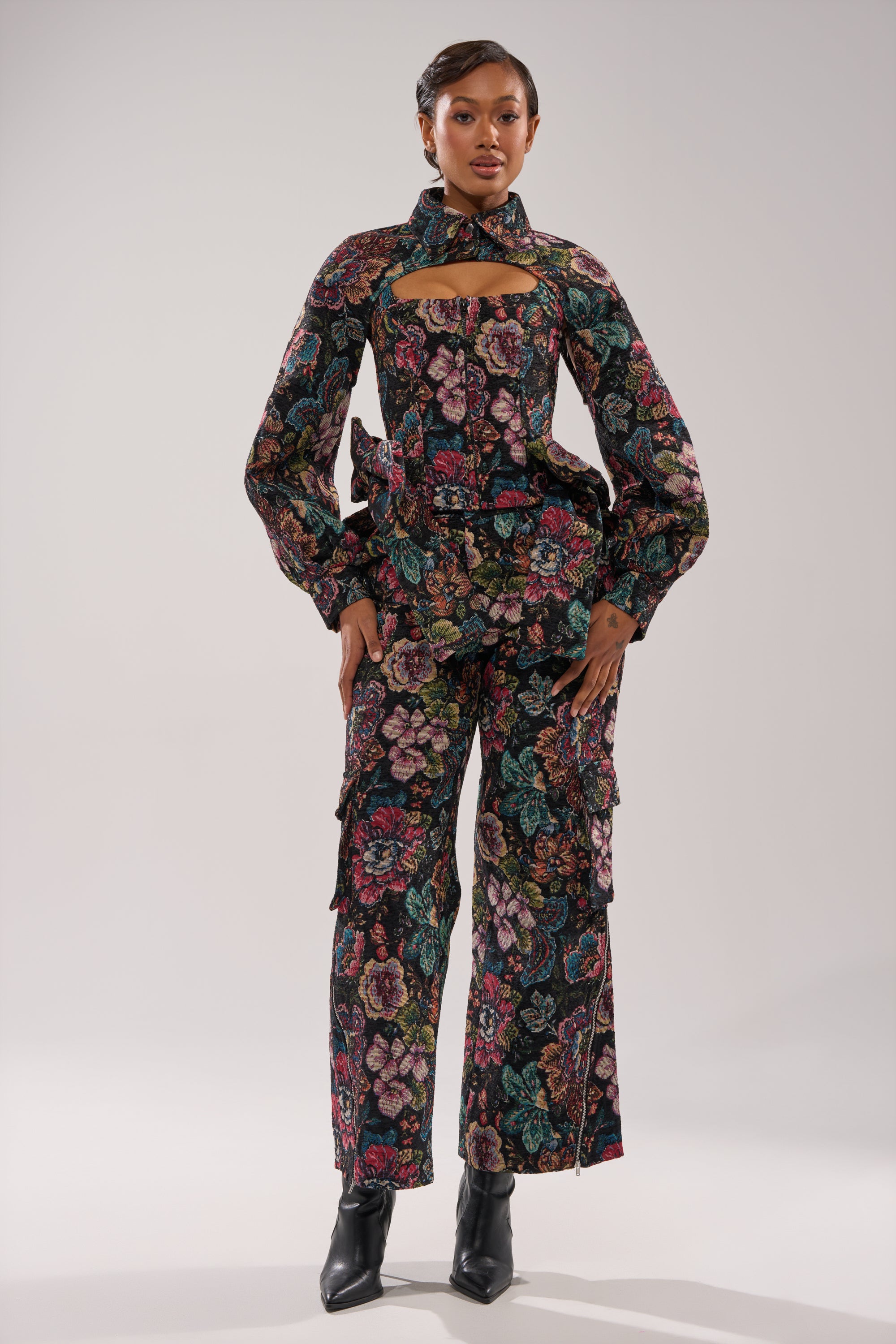 A woman poses in a colorful floral pantsuit and black ankle boots, her look elevated by the PRETTY LITTLE LADY BROCADE CORSET with cutout detail at the chest, all set against a plain light background. 5