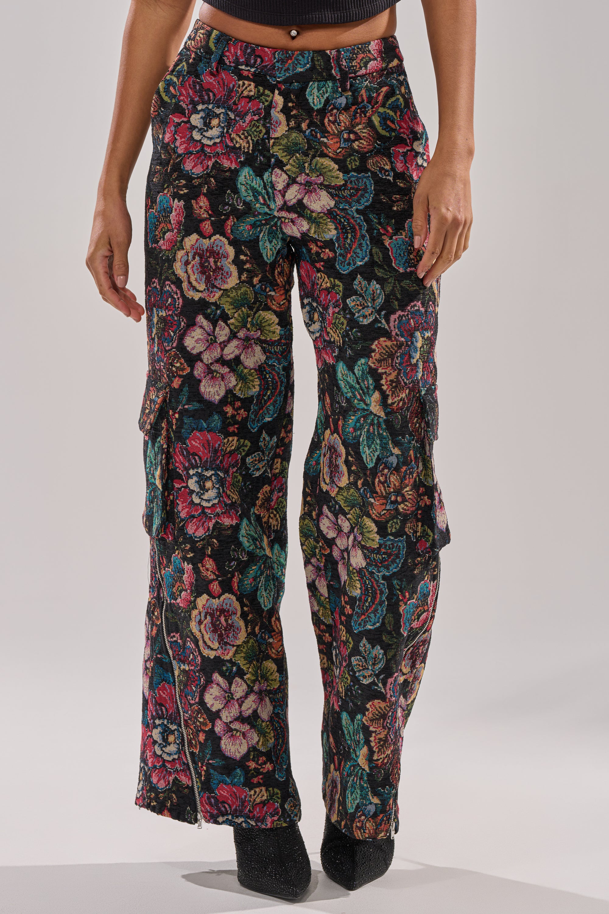 A person wears PRETTY LITTLE LADY BROCADE TROUSER—high-rise, wide-leg pants with a colorful floral pattern—paired with black pointed-toe shoes, standing on a white background for a chic, modern look.