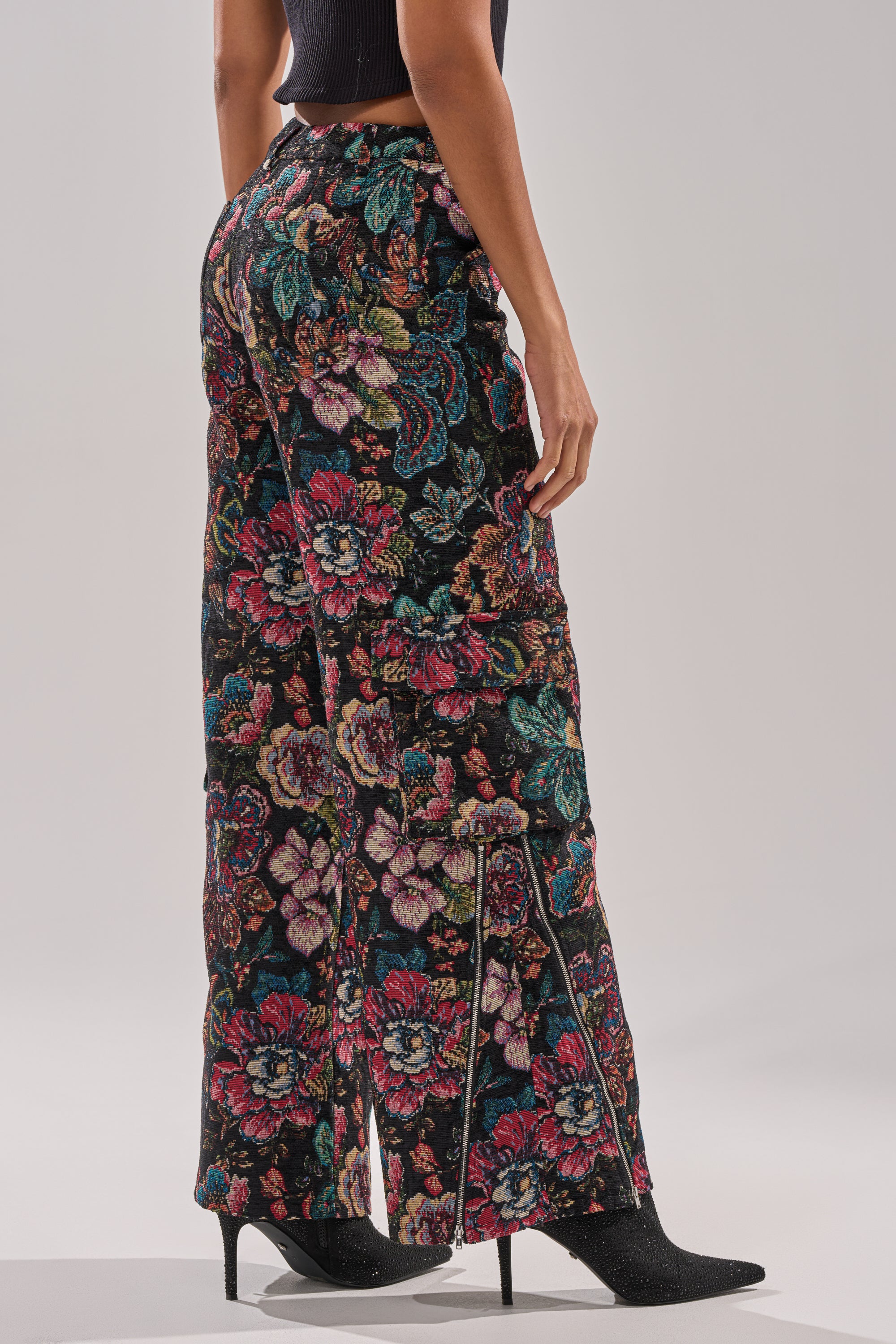 Wearing the PRETTY LITTLE LADY BROCADE TROUSER with vibrant floral patterns, a person pairs it with a black crop top and pointed-toe heeled boots, standing against a plain light backdrop.