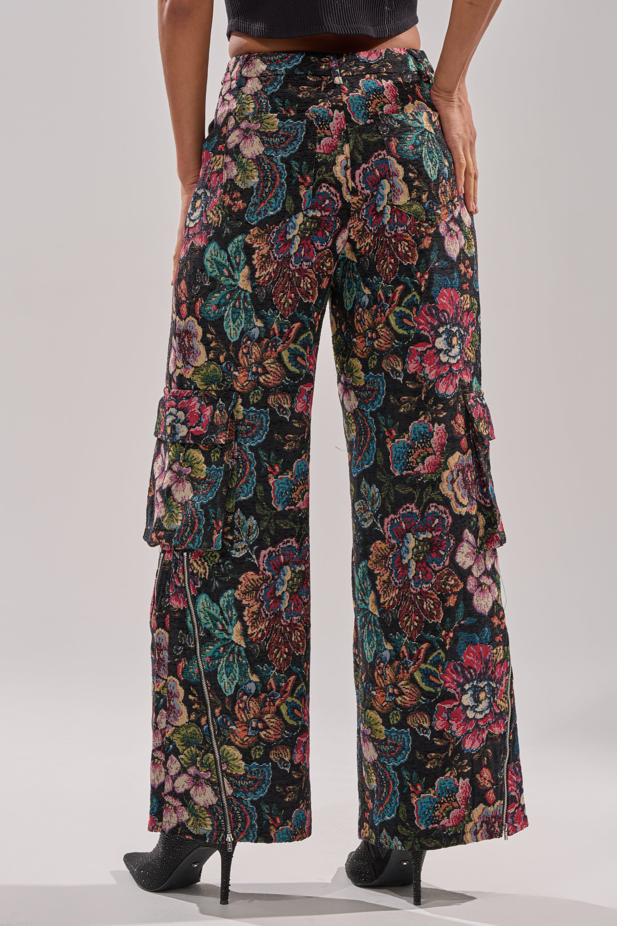 A person stands facing away, wearing PRETTY LITTLE LADY BROCADE TROUSER—black wide-leg trousers with a colorful floral pattern and side cargo pockets—paired with black pointed-toe heels.
