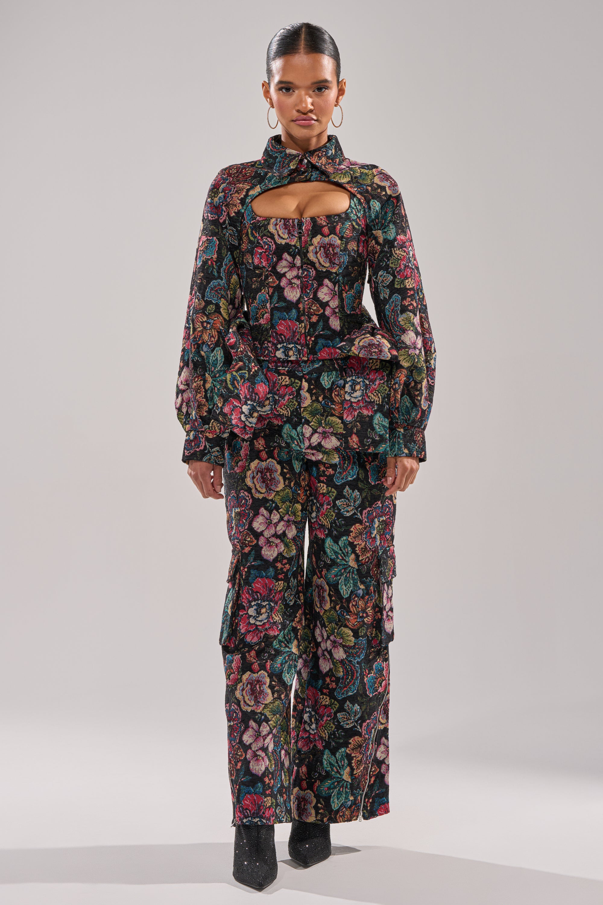 A woman faces forward in the PRETTY LITTLE LADY BROCADE TROUSER set, featuring a floral design, chest cutout, and wide-leg fit. Her hair is pulled back; she wears large hoops and black pointed-toe shoes against a plain background.