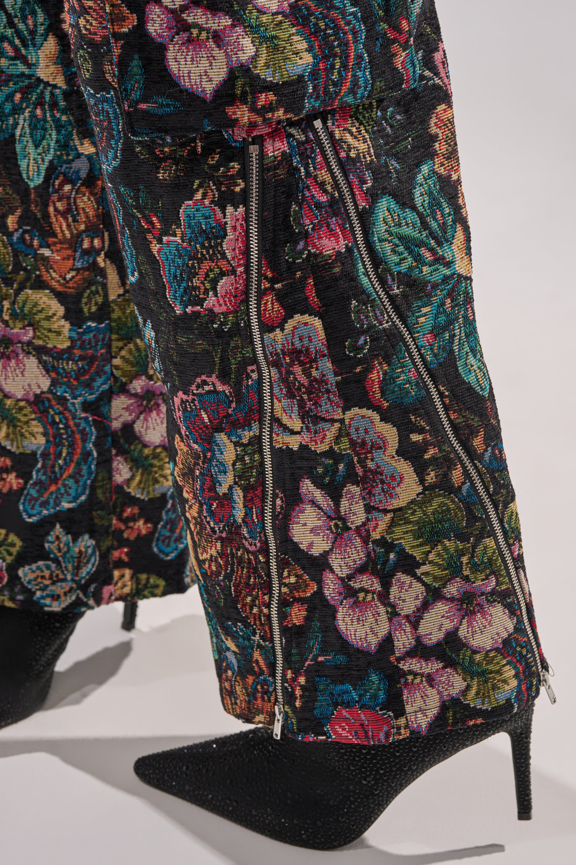 A close-up of legs in PRETTY LITTLE LADY BROCADE TROUSER featuring a colorful floral pattern and visible zippers, paired with pointed black high-heeled shoes.